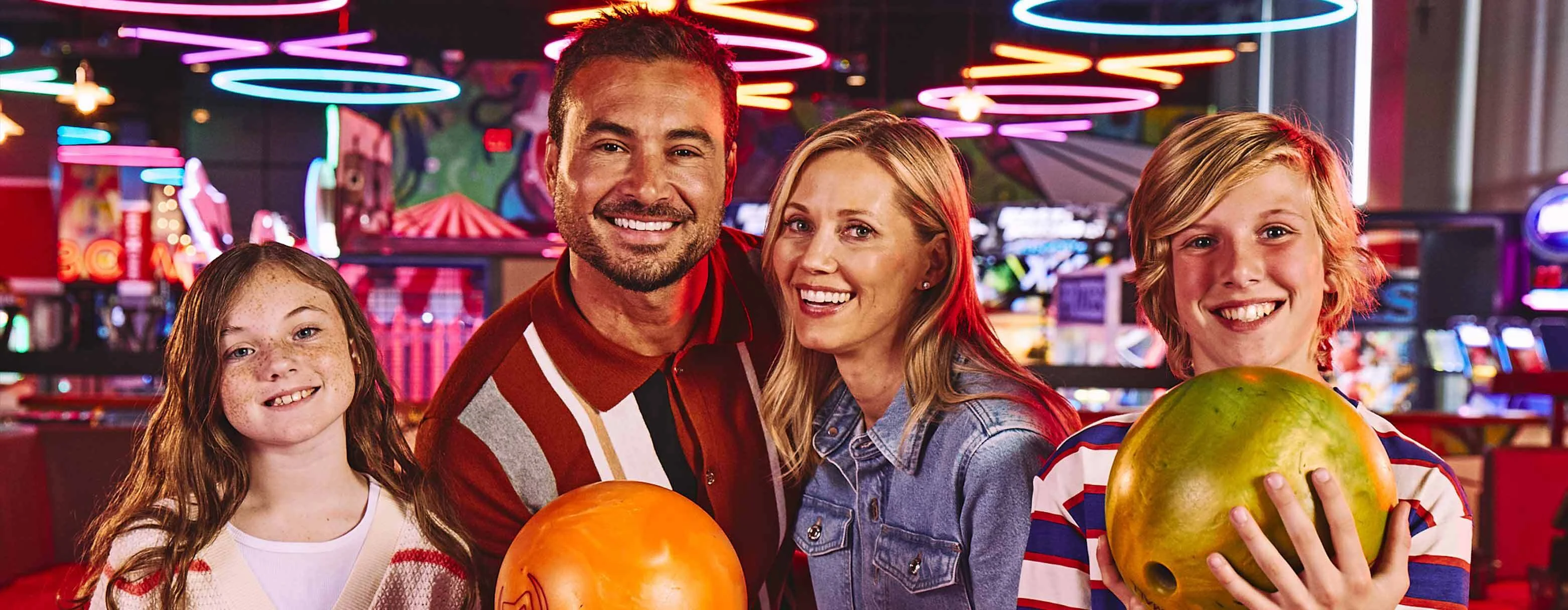 Family of four with bowling balls and colorful arcade in the background.