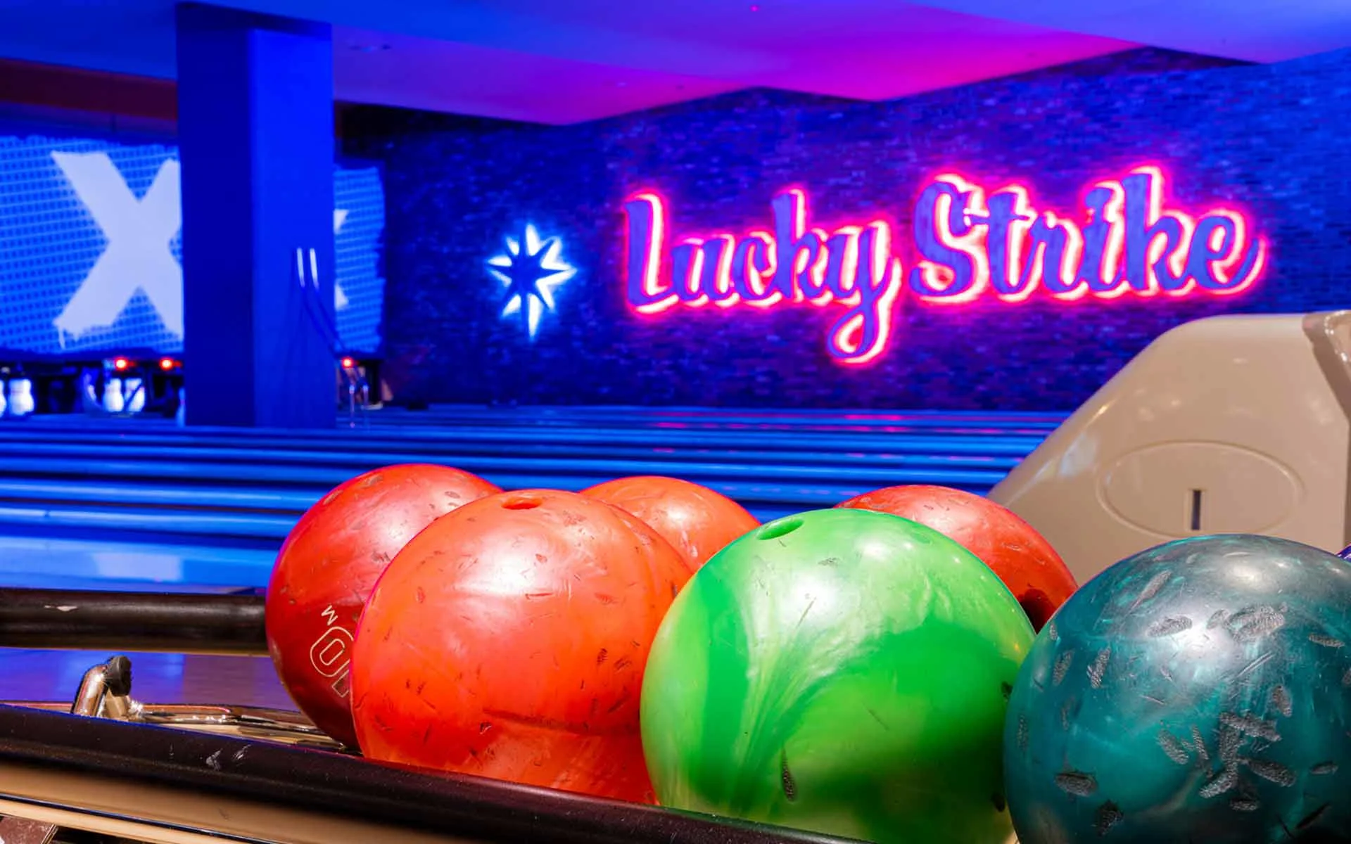 Colorful bowling balls on a rack with neon Lucky Strike signage glowing in the background inside a contemporary bowling alley.