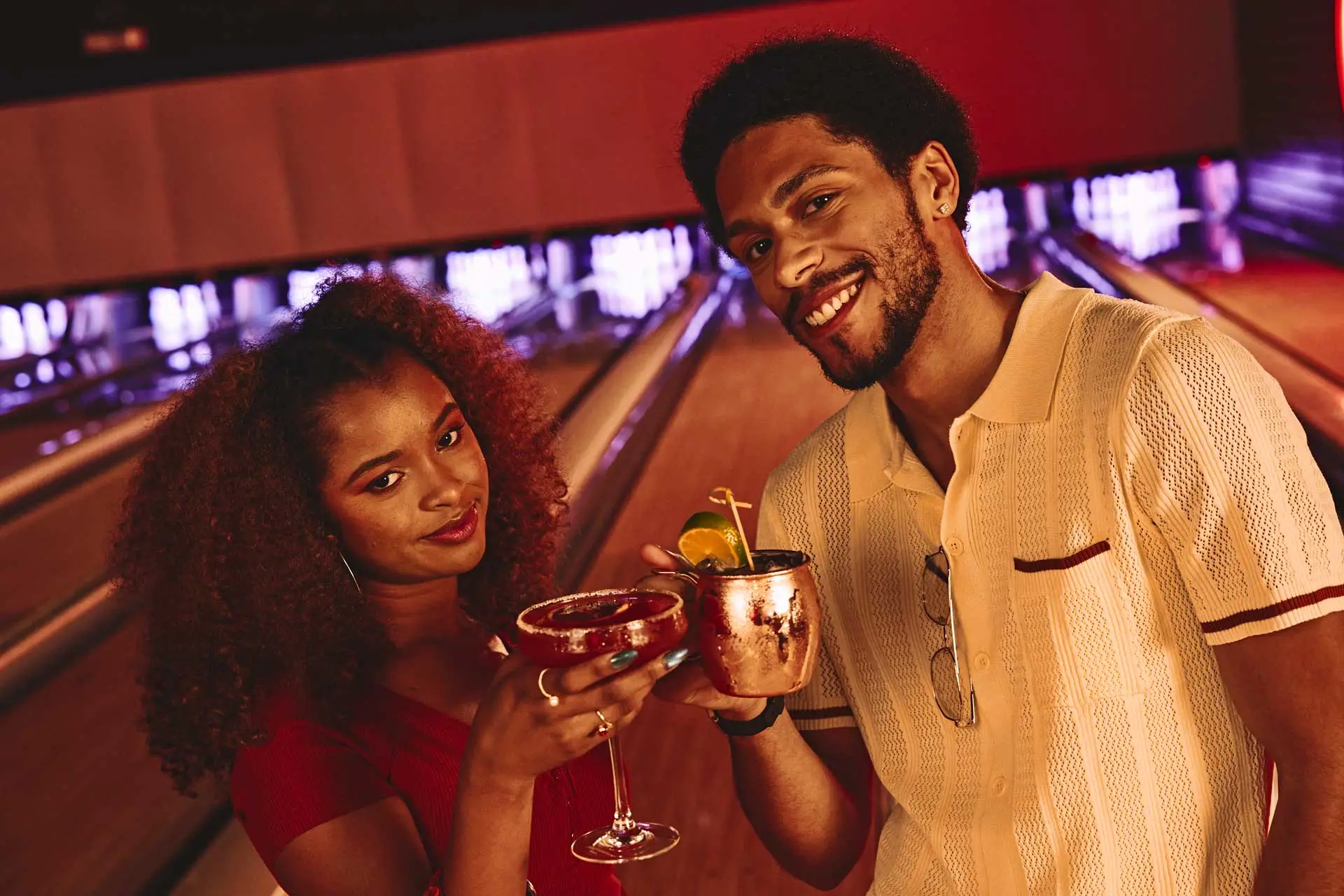 Two adults holding cocktails and smiling beside glowing bowling lanes at an upscale bowling alley with warm neon nightlife lighting.