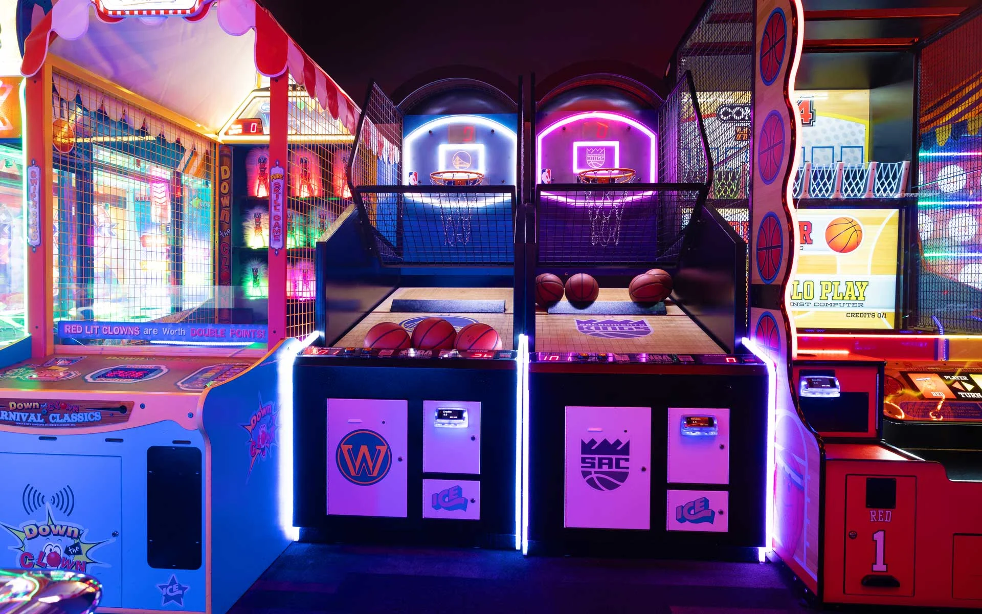 Colorful arcade area with glowing basketball games and carnival-style machines, lit in bright neon pink, blue, and purple.