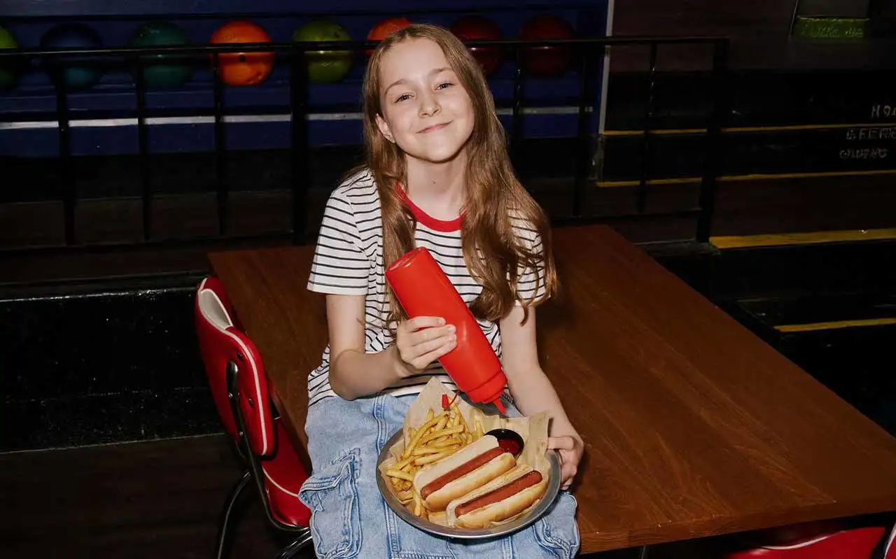 A smiling girl holds a plate of hot dogs and fries with a ketchup bottle at her AMF table by the ball rack.