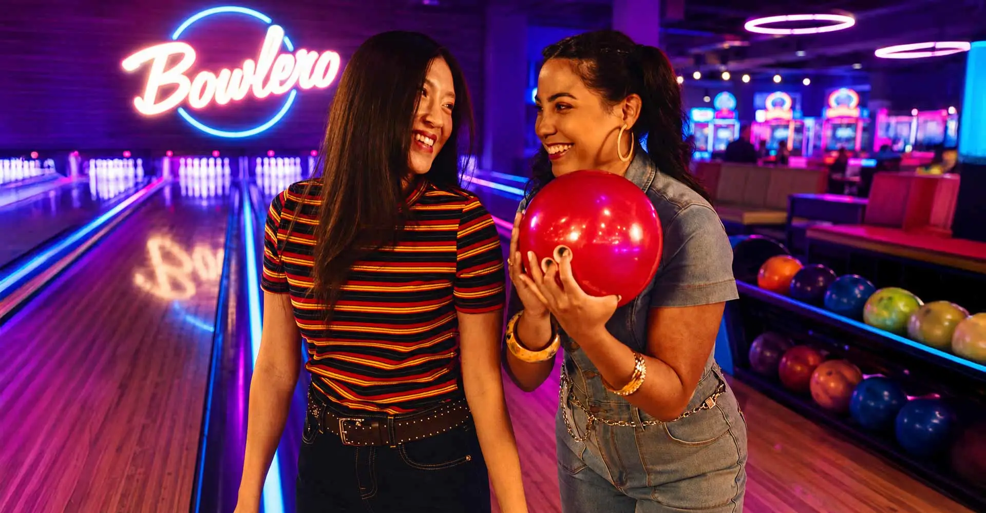 Two women laughing with a red bowling ball inside a neon-lit Bowlero bowling alley with arcade games and colorful lanes.