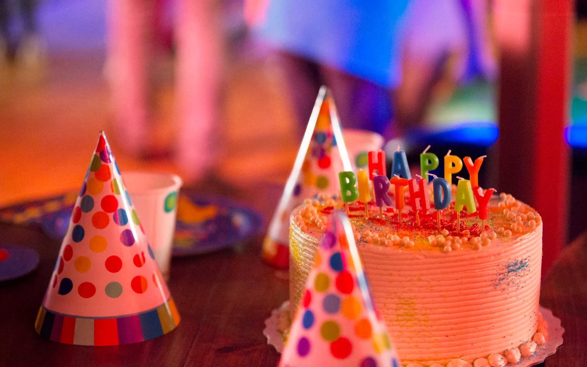 Colorful birthday cake with “Happy Birthday” candles surrounded by party hats on a table in a vibrant, festive setting.