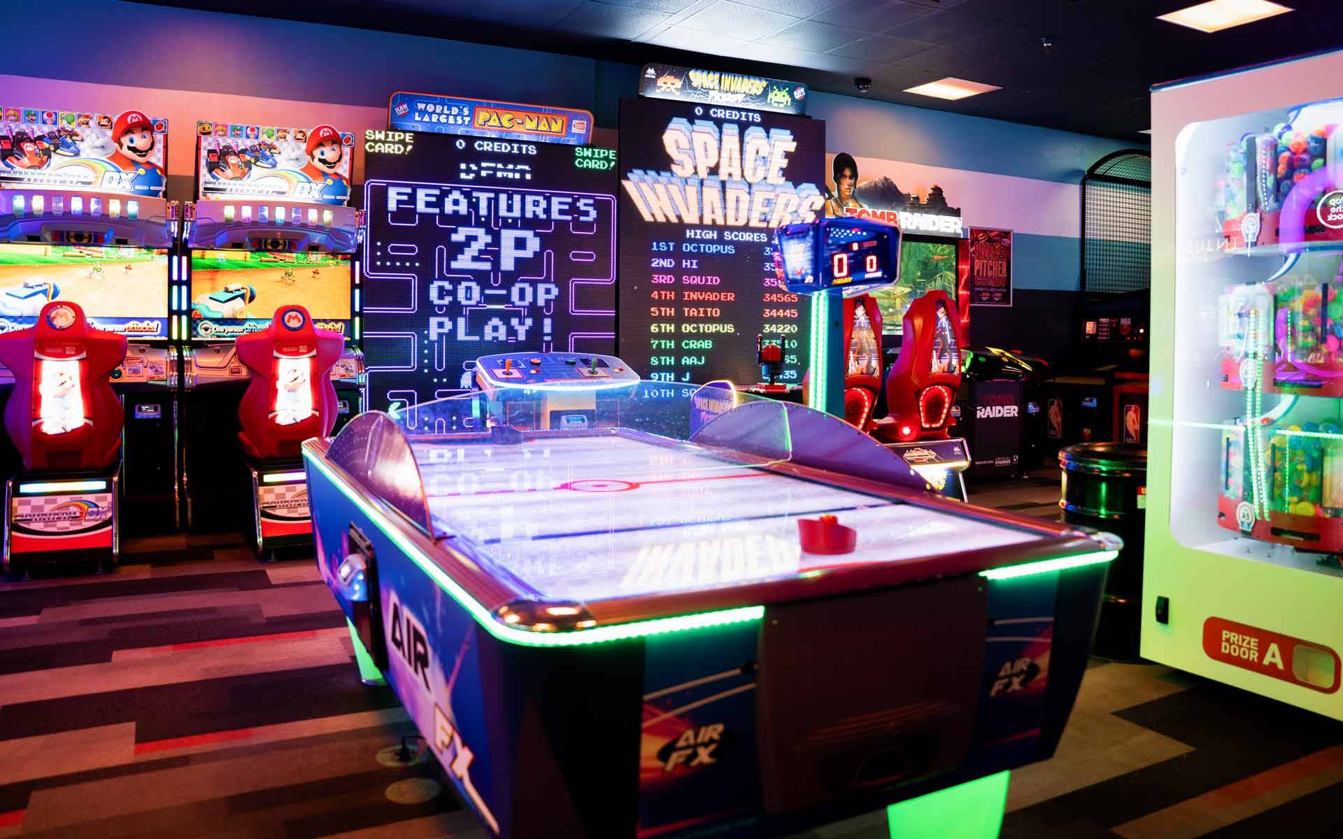 Vibrant arcade area with air hockey table, Space Invaders game wall, racing simulators, and prize machines inside a Lucky Strike entertainment center.