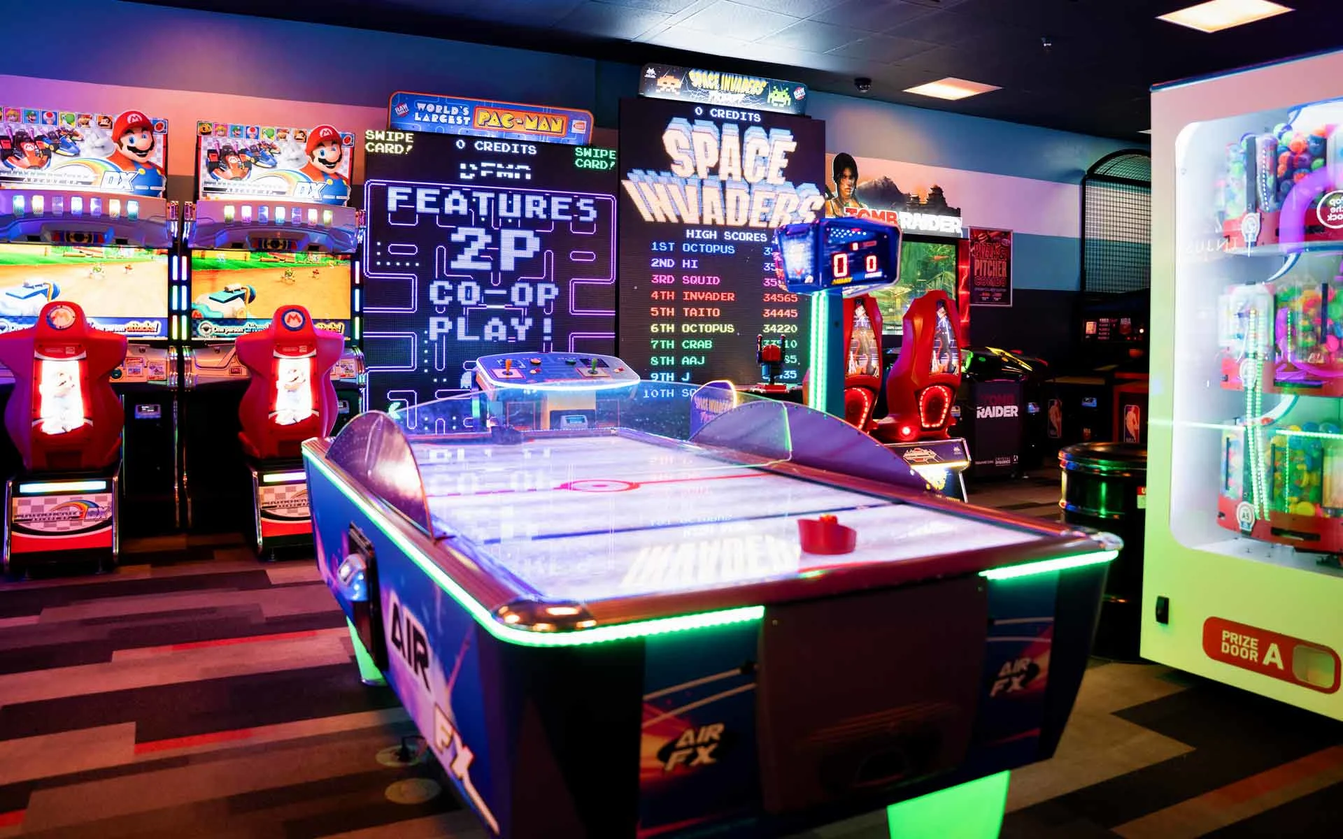 Vibrant arcade area with air hockey table, Space Invaders game wall, racing simulators, and prize machines inside a Lucky Strike entertainment center.