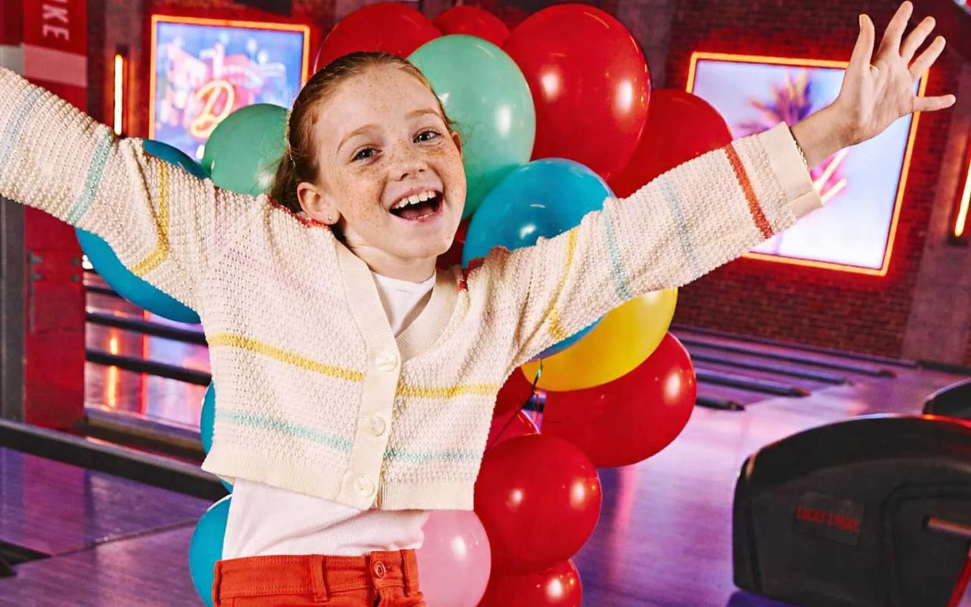 Smiling young girl sitting on a bowling ball return with arms raised, surrounded by colorful balloons at a Lucky Strike bowling center.