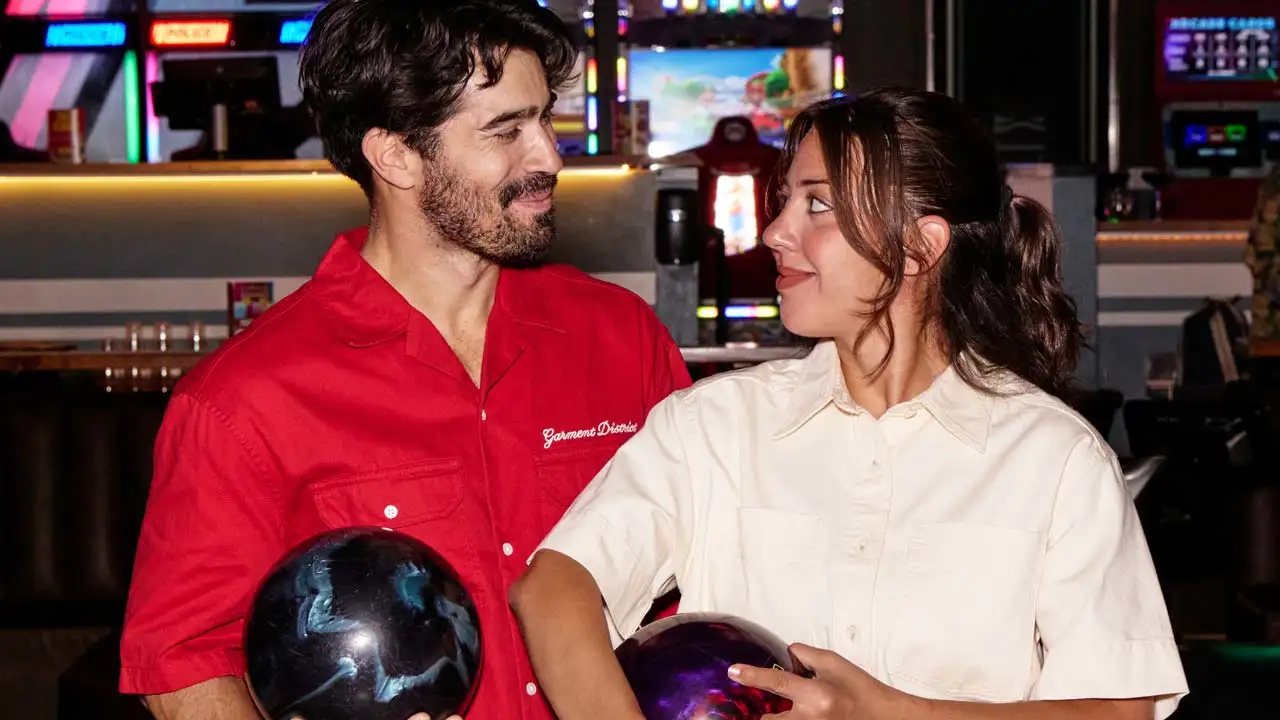 Two people holding bowling balls and smiling at each other inside a bowling entertainment center with arcade games in the background.