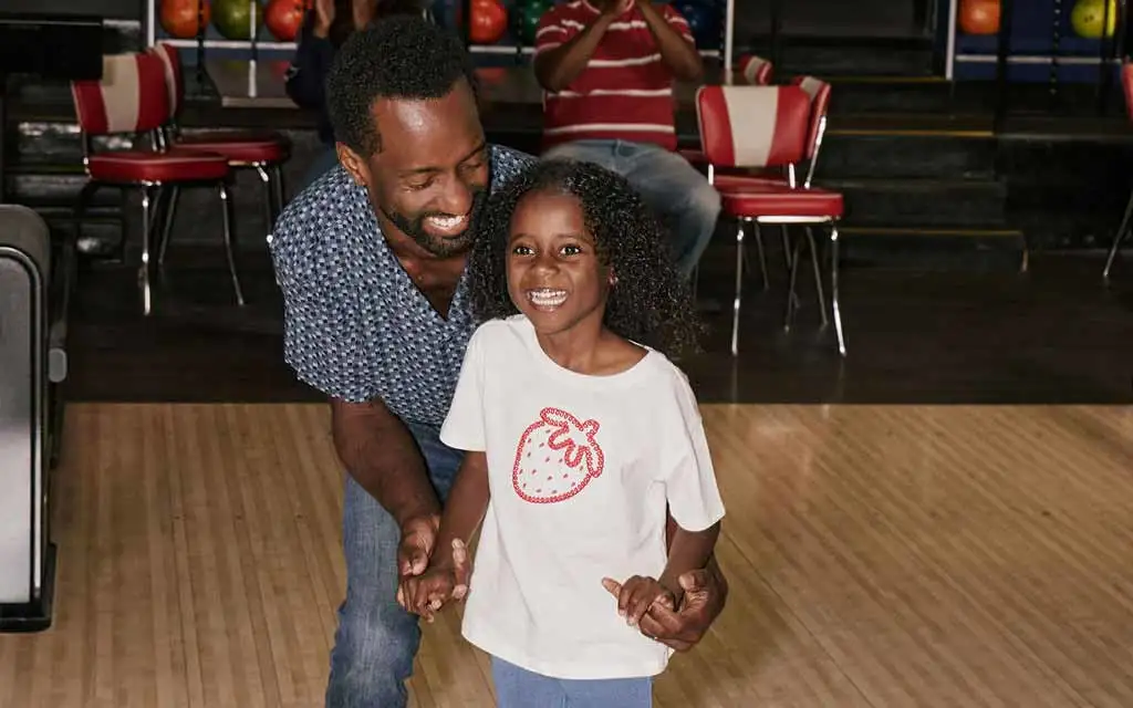 A father and his young daughter laugh together on the approach of an AMF bowling lane.
