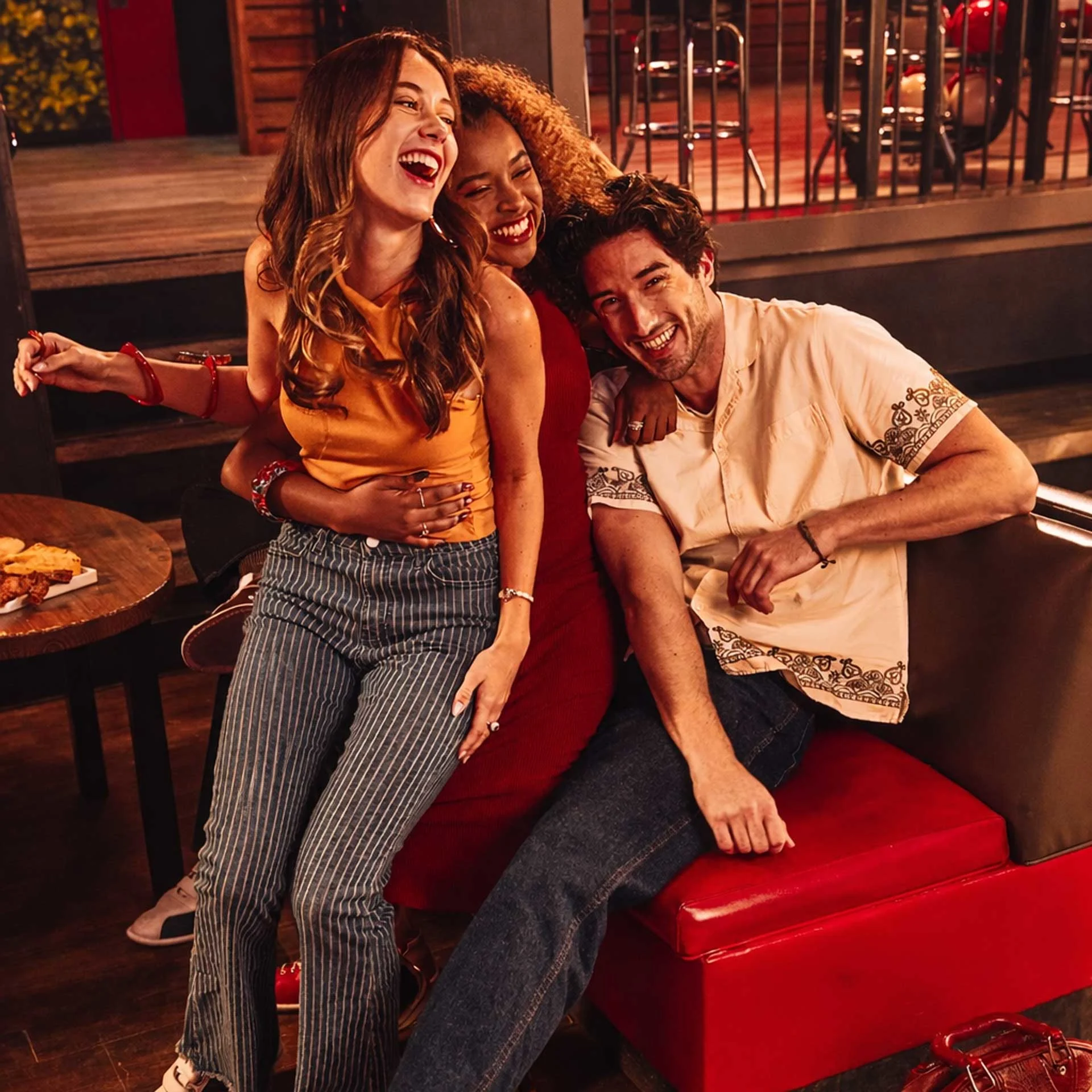 Three friends laughing together in a bowling lounge seating area, with drinks and food on a table nearby.