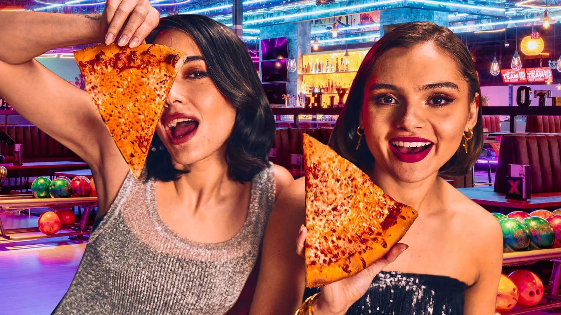 Two ladies holding slices of pizza at a bowling alley.