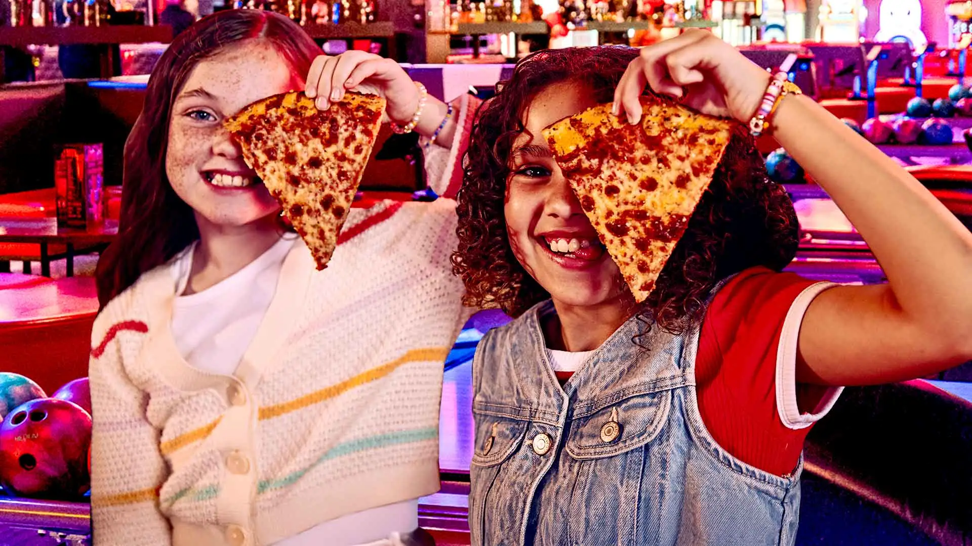Two girls holding slices of pizza at a bowling alley.