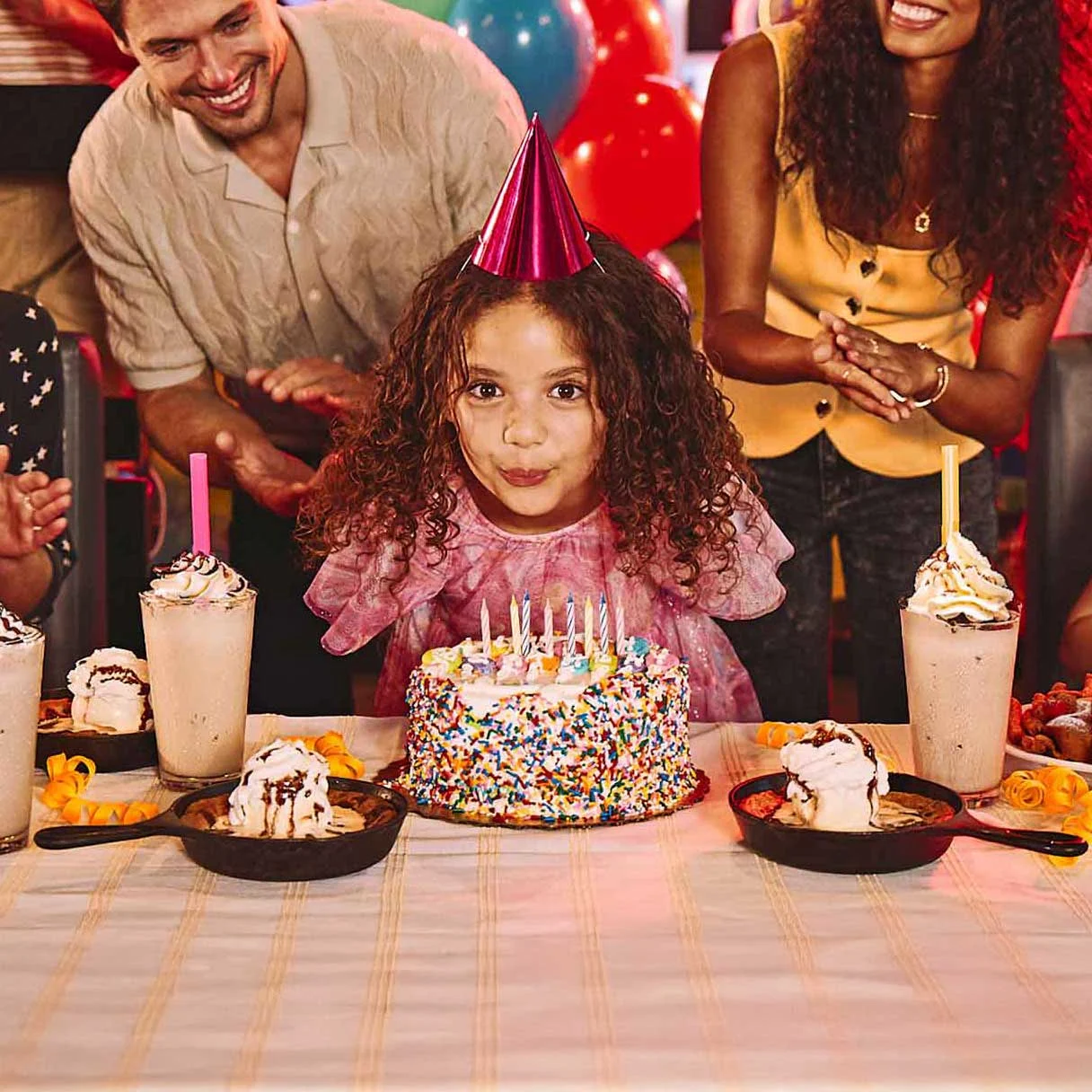 Children’s birthday party at a bowling alley with cake, milkshakes, balloons, and guests clapping in a neon-lit venue.