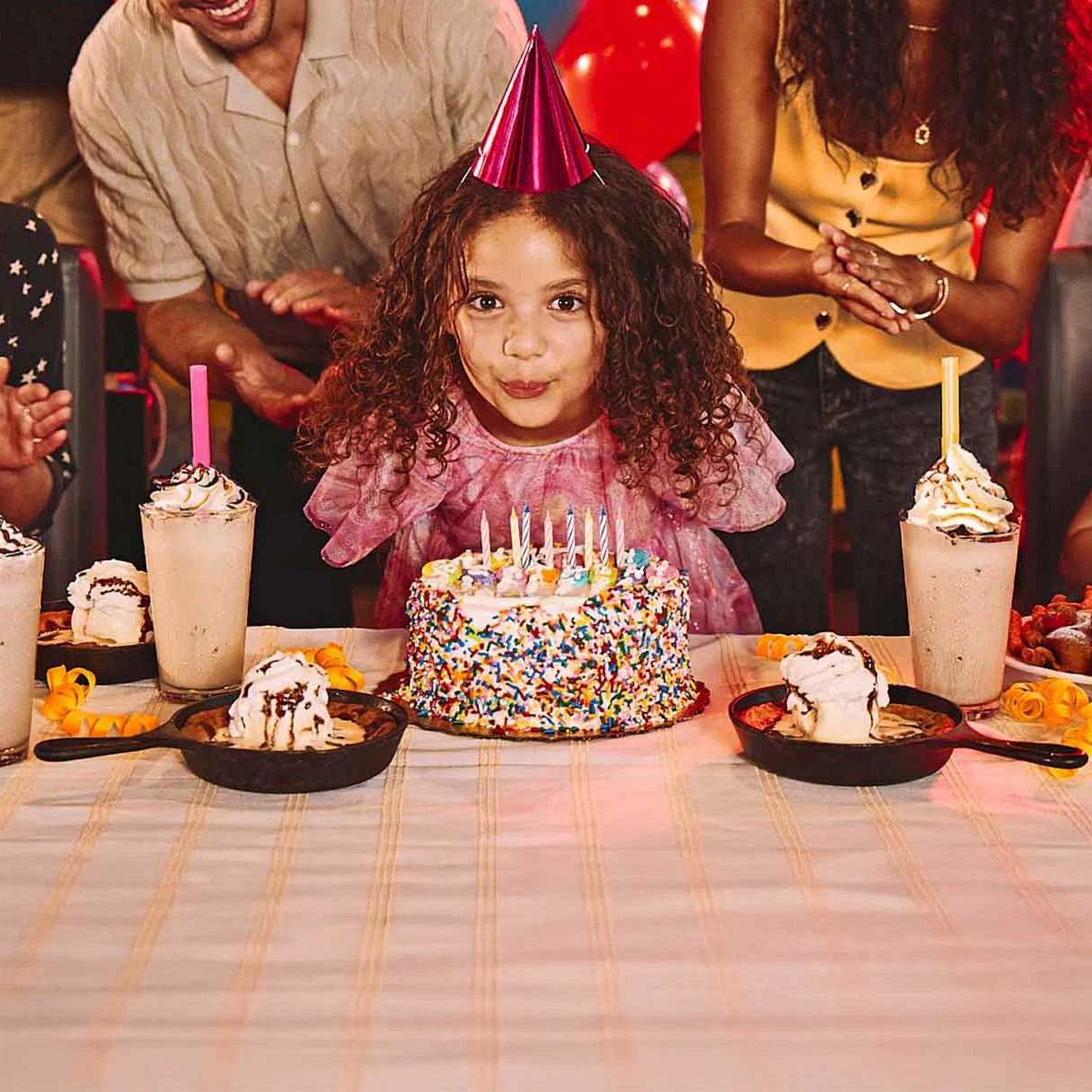 Children’s birthday party at a bowling alley with cake, milkshakes, balloons, and guests clapping in a neon-lit venue.