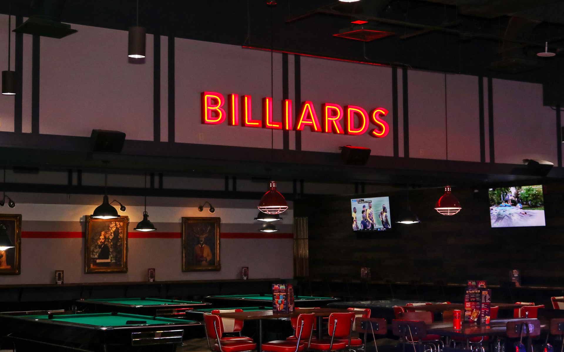 Upscale bowling venue billiards area with pool tables, red seating, and neon “Billiards” sign on the wall