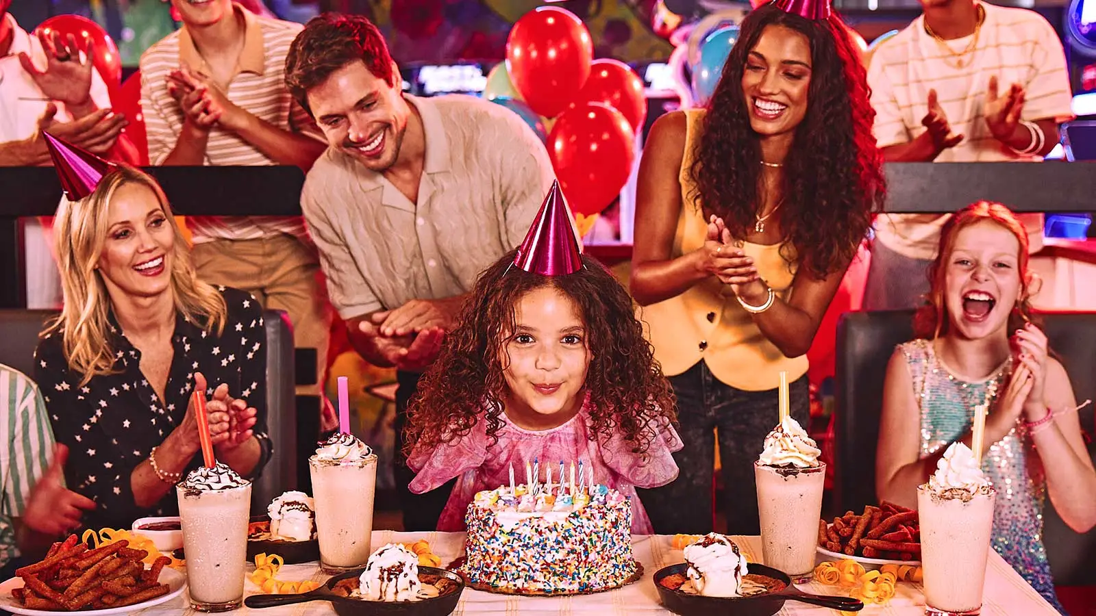 A girl with a birthday cake and a large group of people in the back celebrating her birthday. 