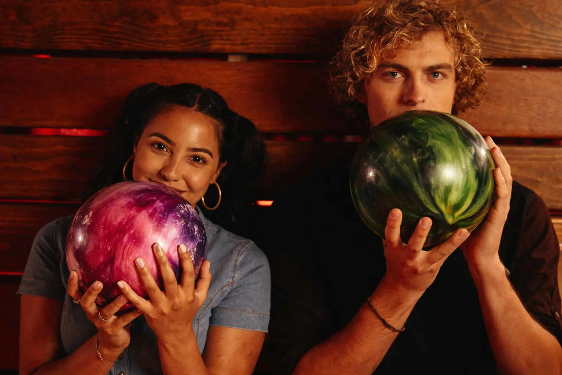 Young couple posing with colorful custom bowling balls—pink-purple and green—held up near their faces against a warm wooden backdrop.