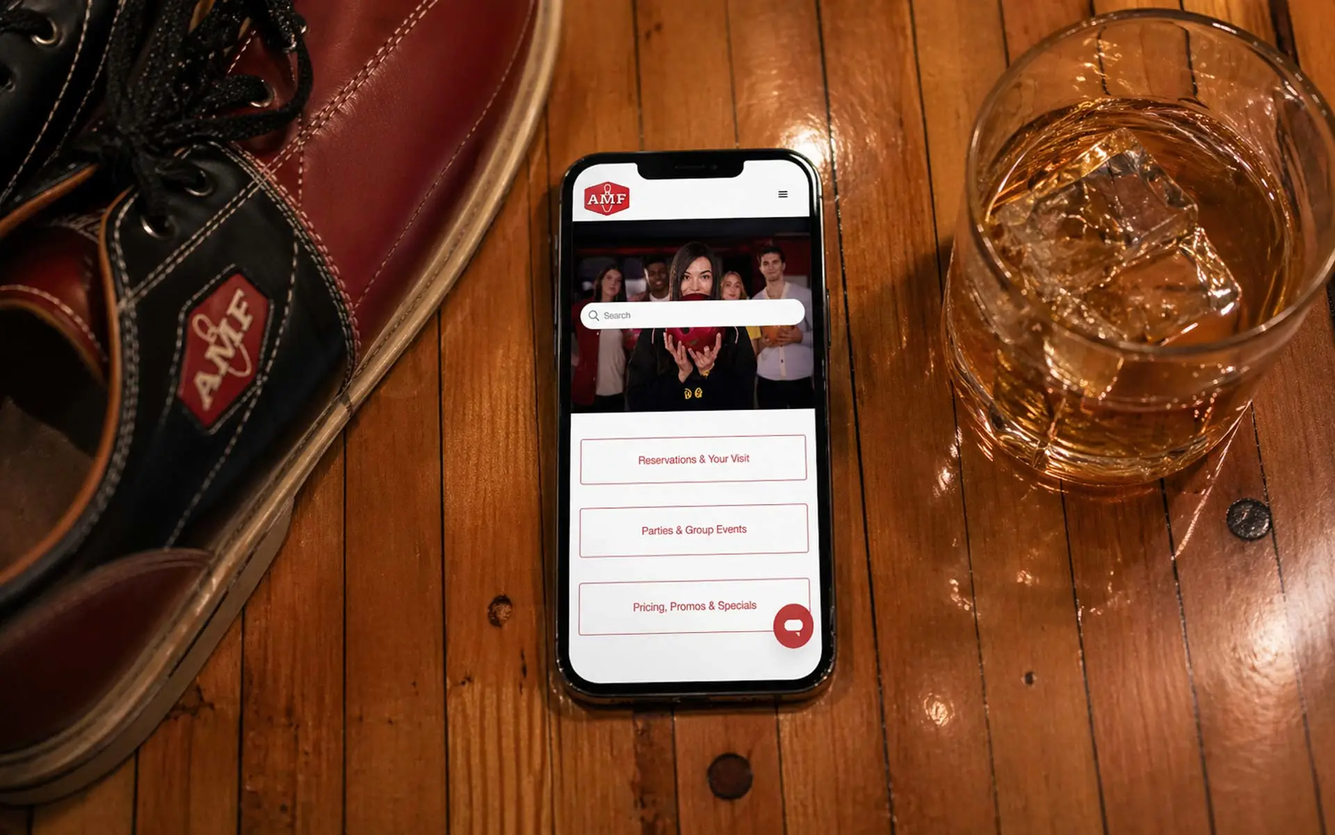 Modern smartphone displaying AMF booking interface on a glossy bowling lane table, with vintage rental shoes and a whiskey glass under warm ambient lighting