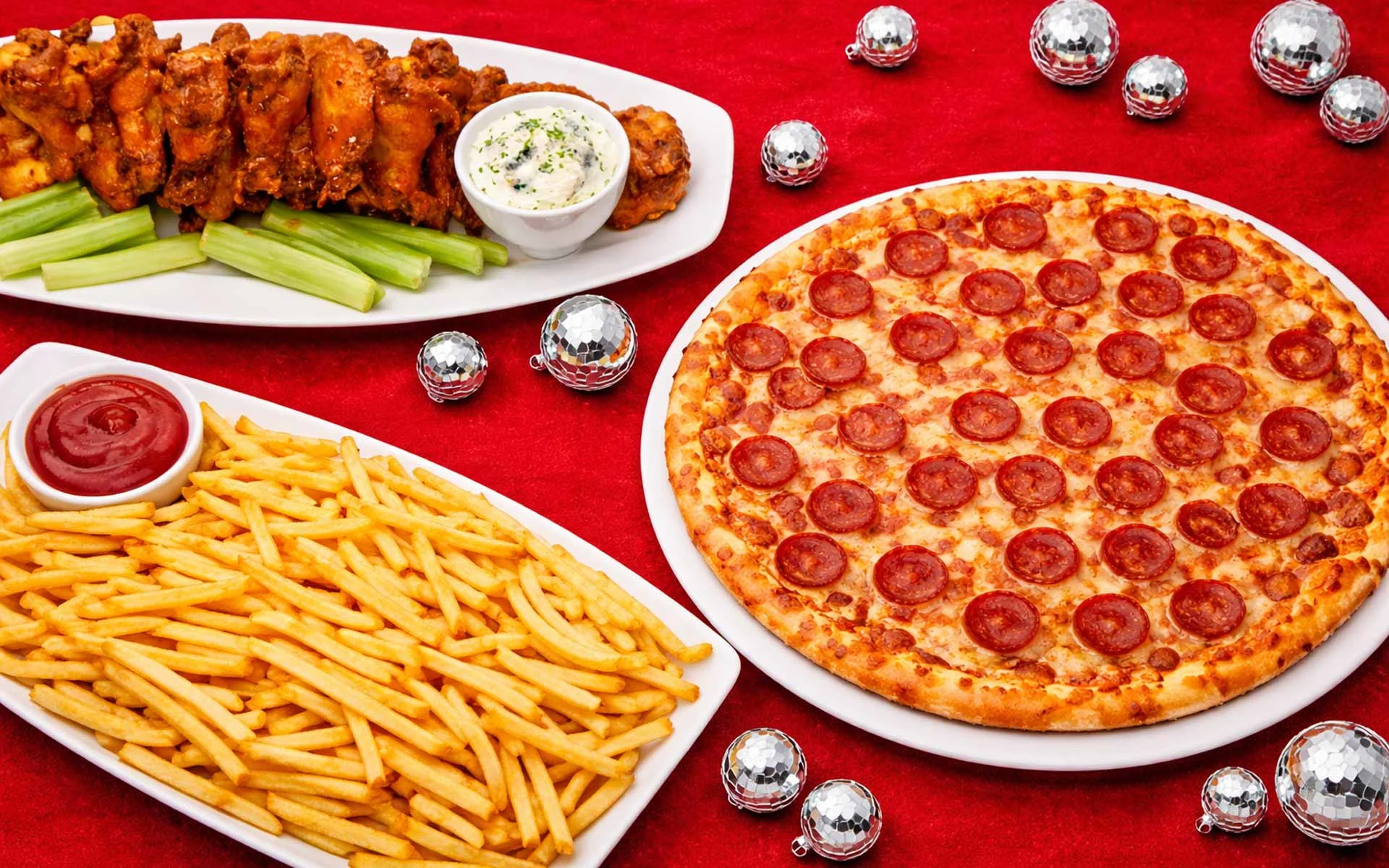 Party food spread on a red table featuring buffalo wings with celery and blue cheese dip, thin-cut French fries with ketchup, and a large pepperoni pizza on a white plate, accented by scattered disco balls.