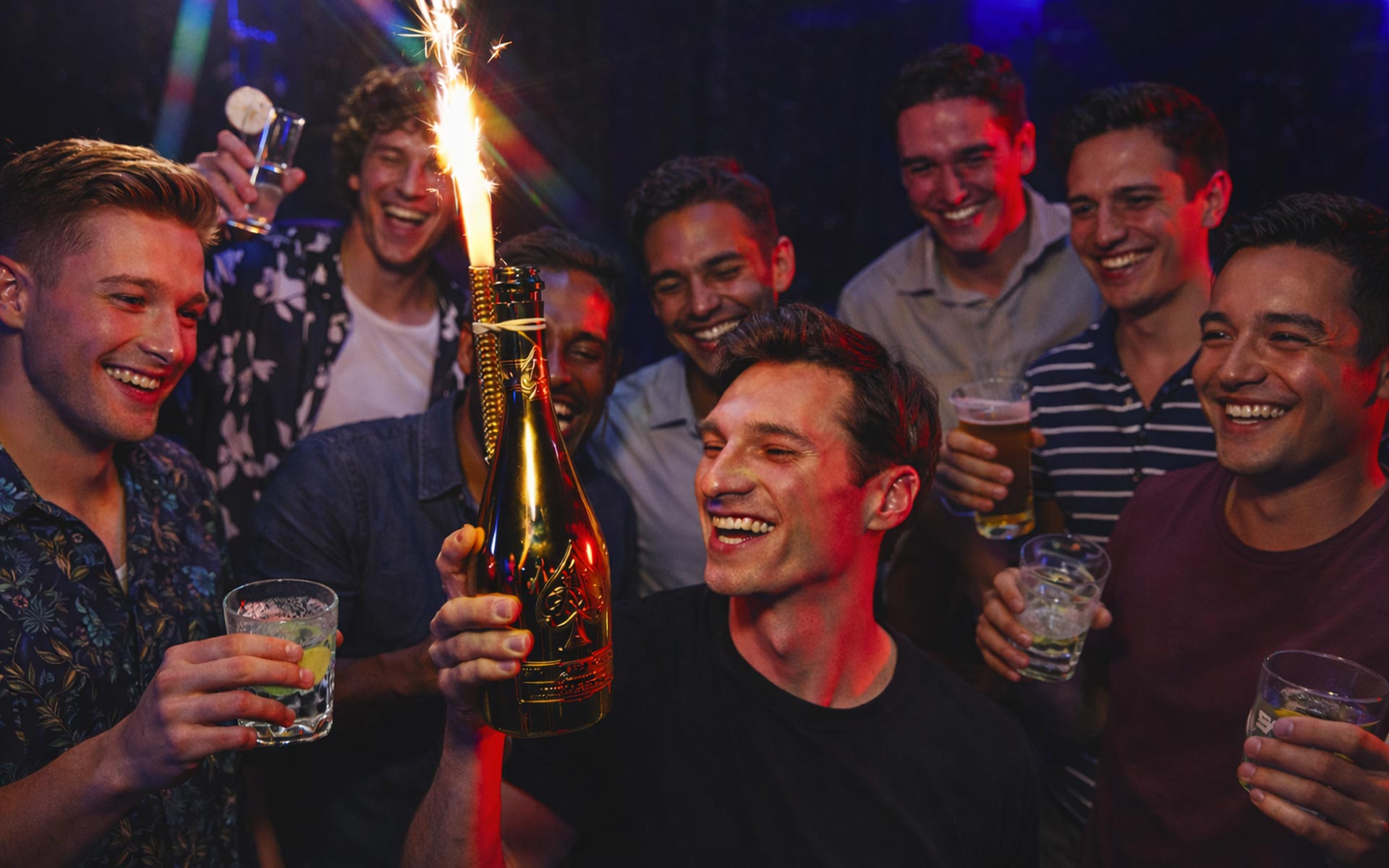 Group of young men celebrating a bachelor party in a nightclub, smiling and holding drinks as one man holds a champagne bottle with a sparkler, surrounded by colorful lighting and a lively atmosphere.