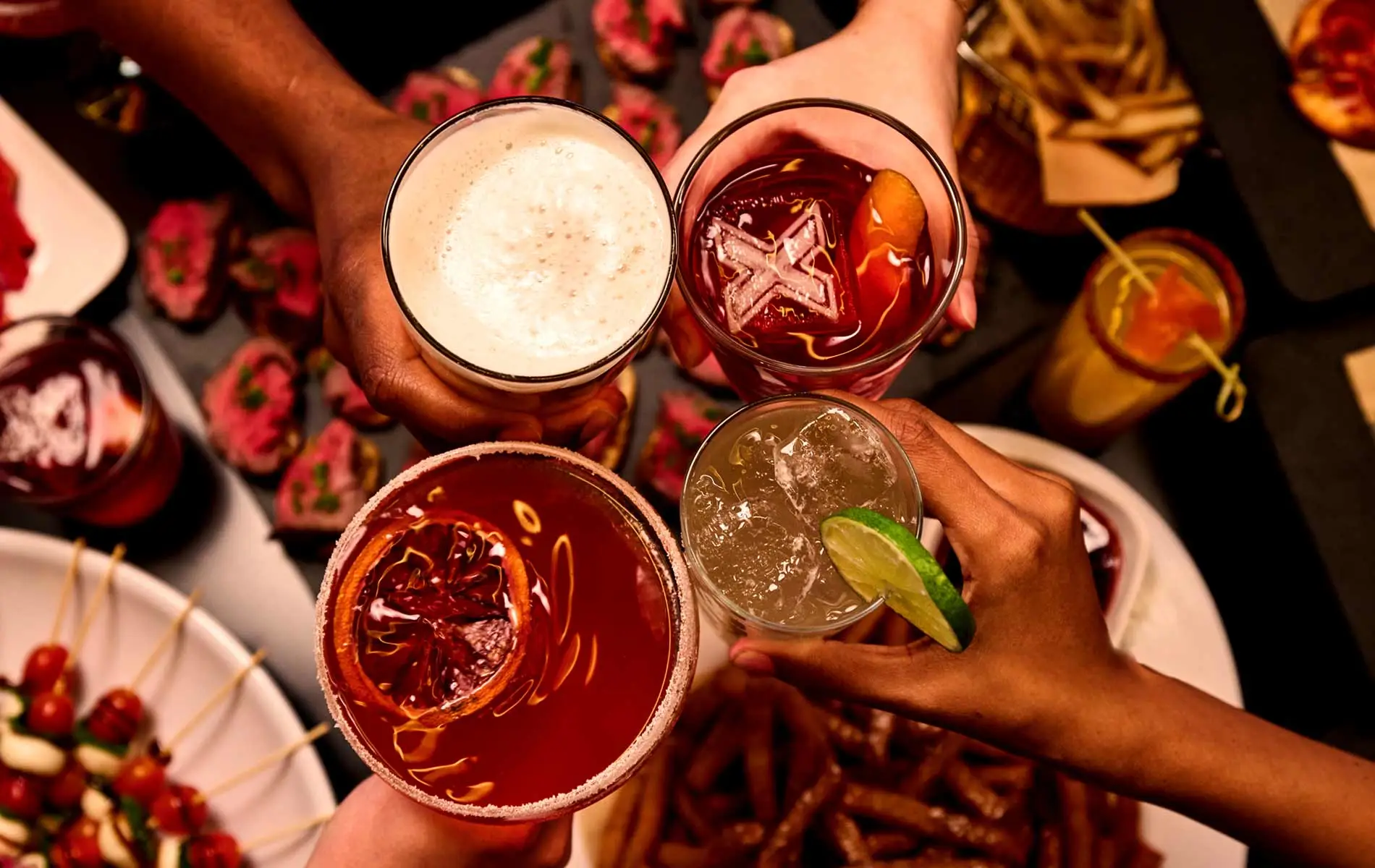 four people holding drinks over an event menu spread