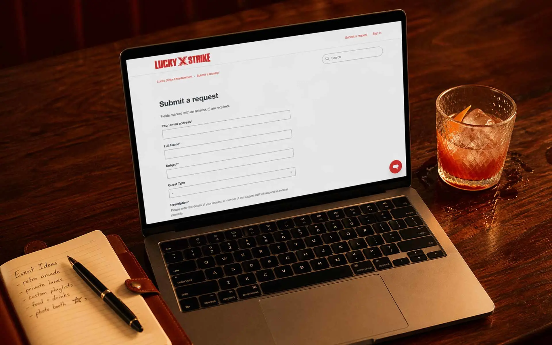 Overhead flat-lay of a laptop showing Lucky Strike “Submit a Request” contact form on a wood bar, with a craft cocktail and leather notebook in a modern bowling lounge setting.