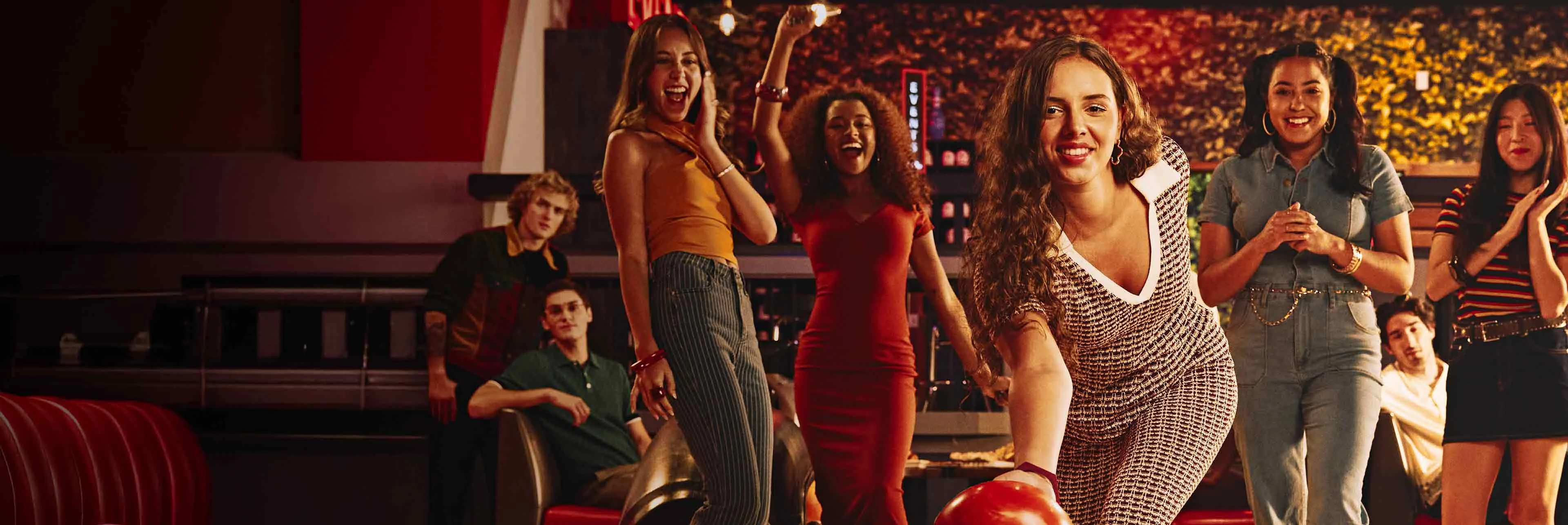 A woman rolls a bowling ball down the lane while a group of friends cheer behind her inside a lively, neon-lit bowling alley filled with people socializing and watching the game.