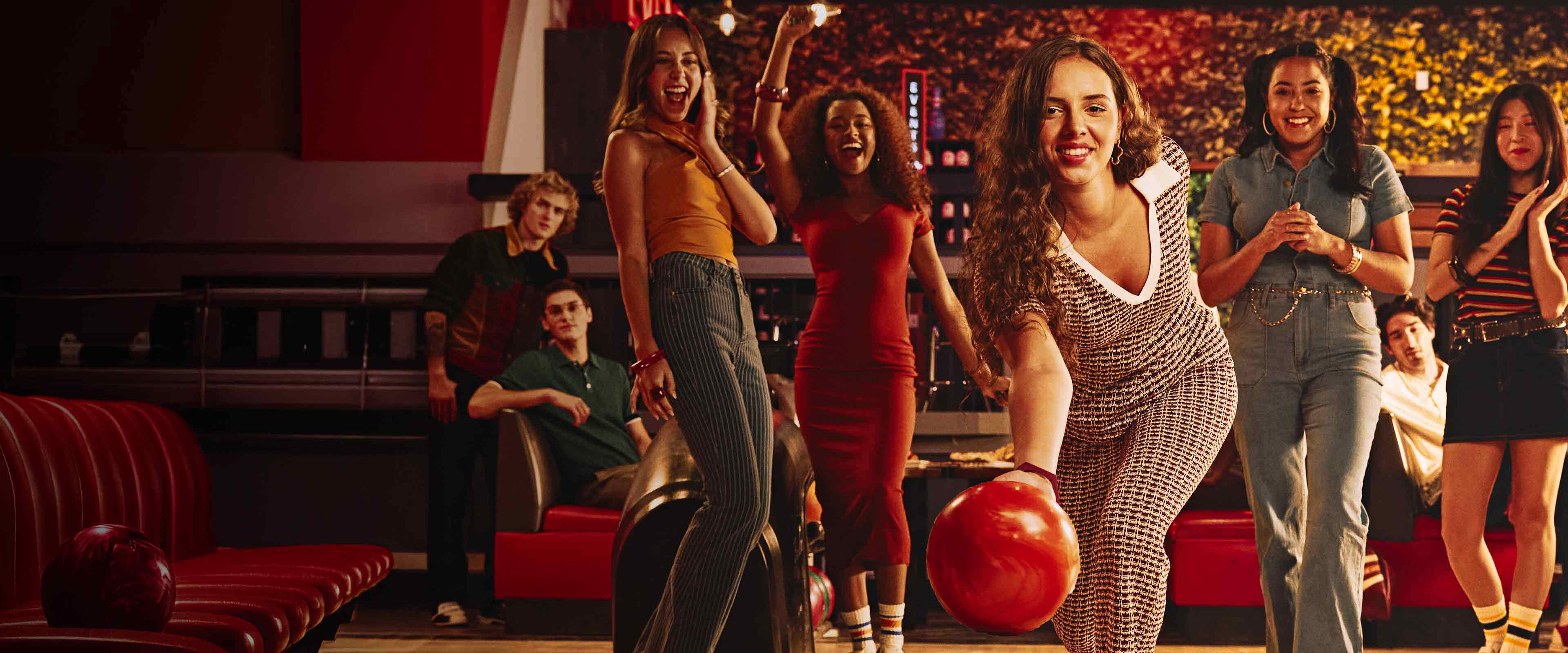 A woman rolls a bowling ball down the lane while a group of friends cheer behind her inside a lively, neon-lit bowling alley filled with people socializing and watching the game.