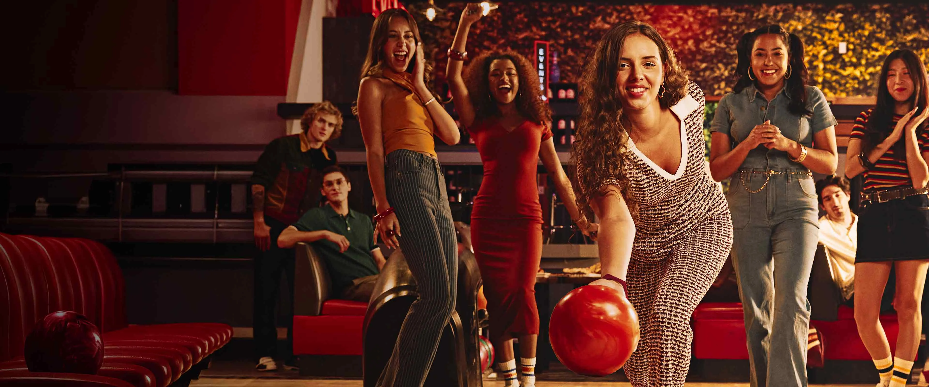 A woman rolls a bowling ball down the lane while a group of friends cheer behind her inside a lively, neon-lit bowling alley filled with people socializing and watching the game.