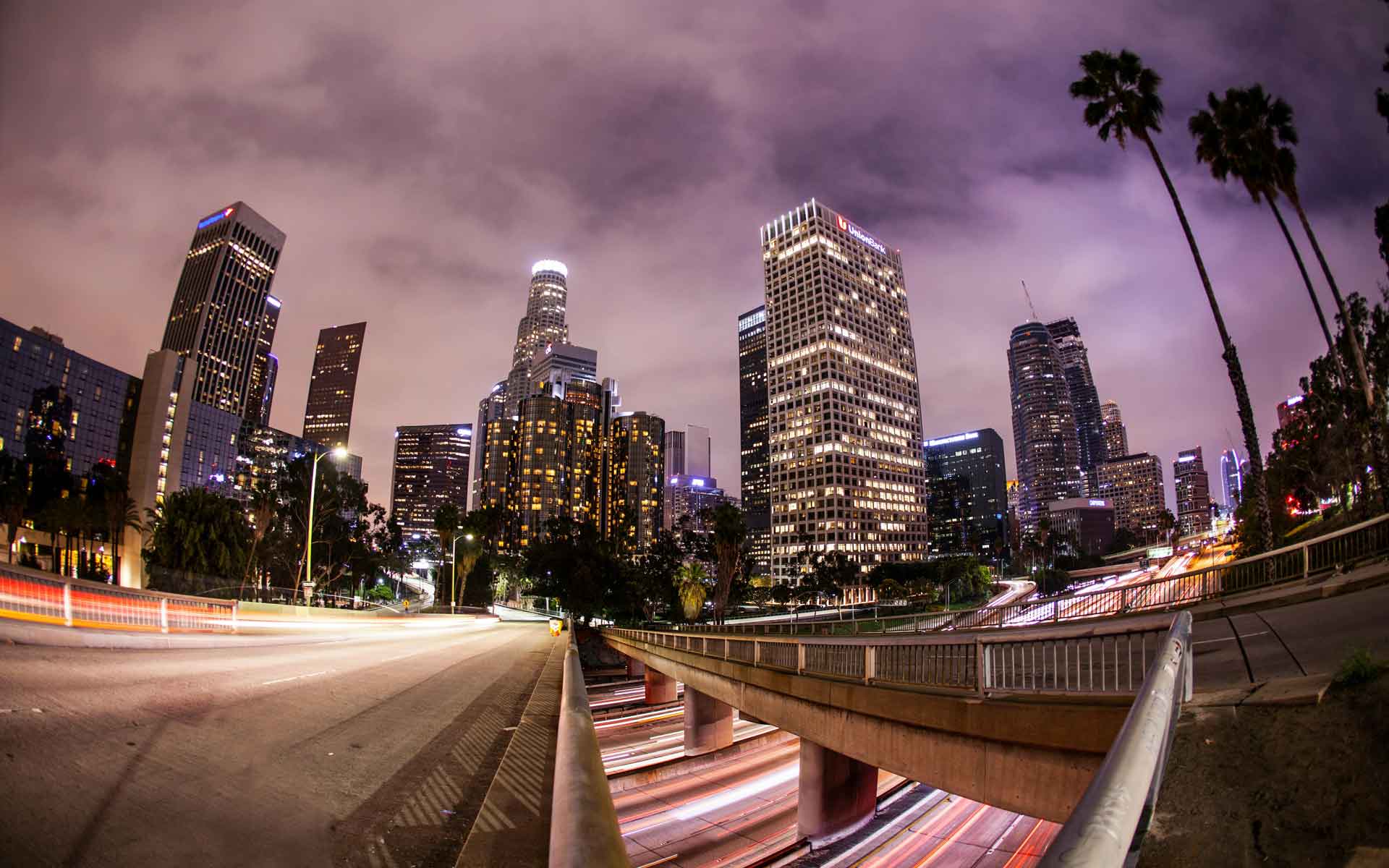Downtown Los Angeles skyline at night with illuminated skyscrapers, palm trees, and freeway light trails.