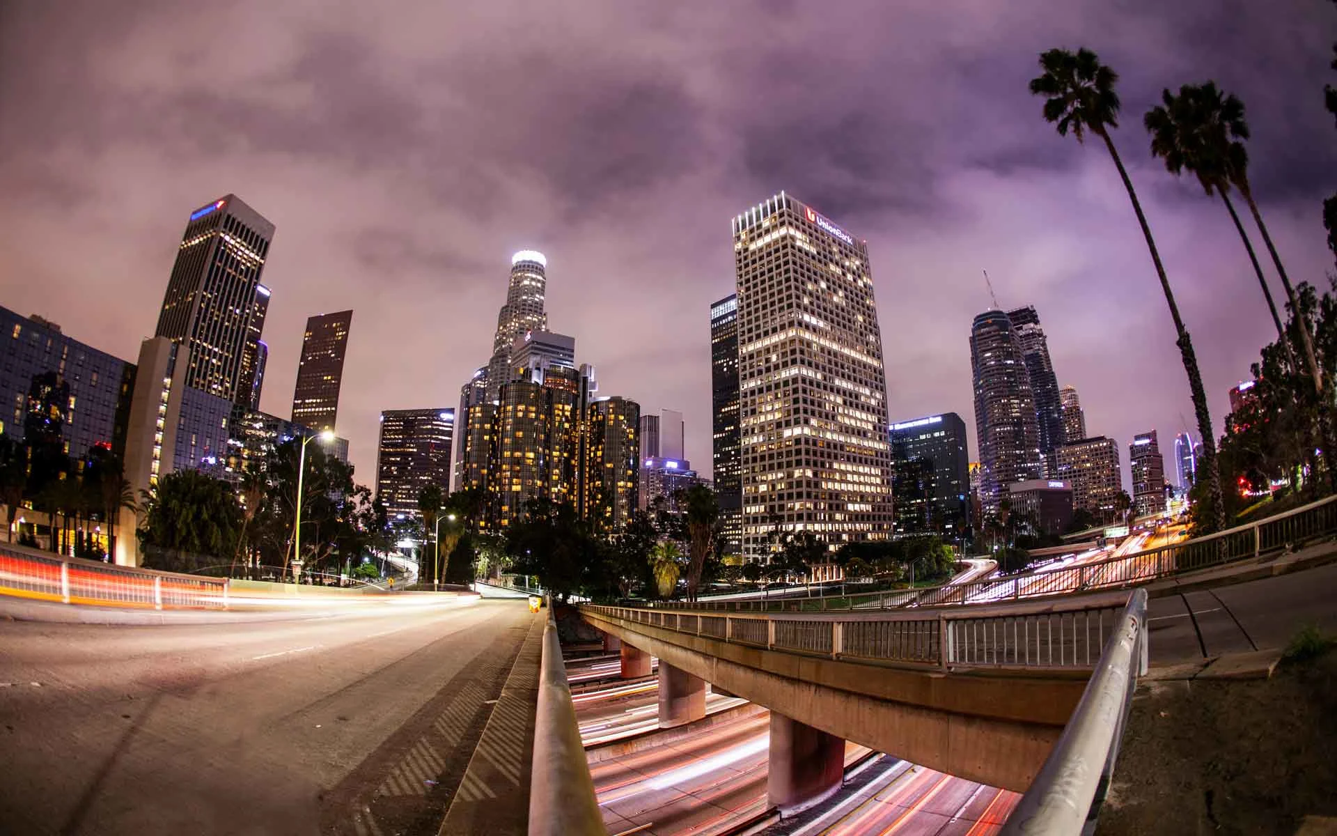 Downtown Los Angeles skyline at night with illuminated skyscrapers, palm trees, and freeway light trails.