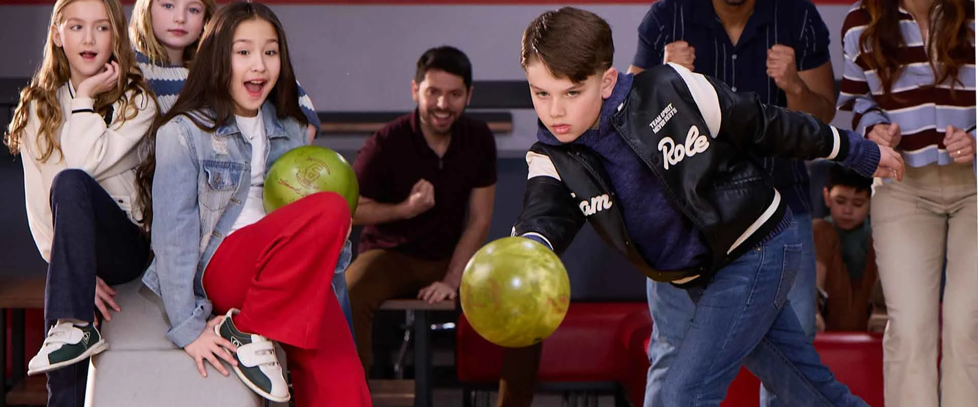 Kids cheering as a boy throws a bowling ball at AMF, with family watching from the seating area.