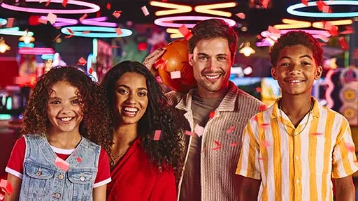 A family smiling together at Lucky Strike with colorful arcade lights in the background, holding a bowling ball and celebrating during a kids’ bowling party.
