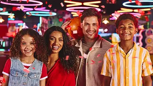 A family smiling together at Lucky Strike with colorful arcade lights in the background, holding a bowling ball and celebrating during a kids’ bowling party.