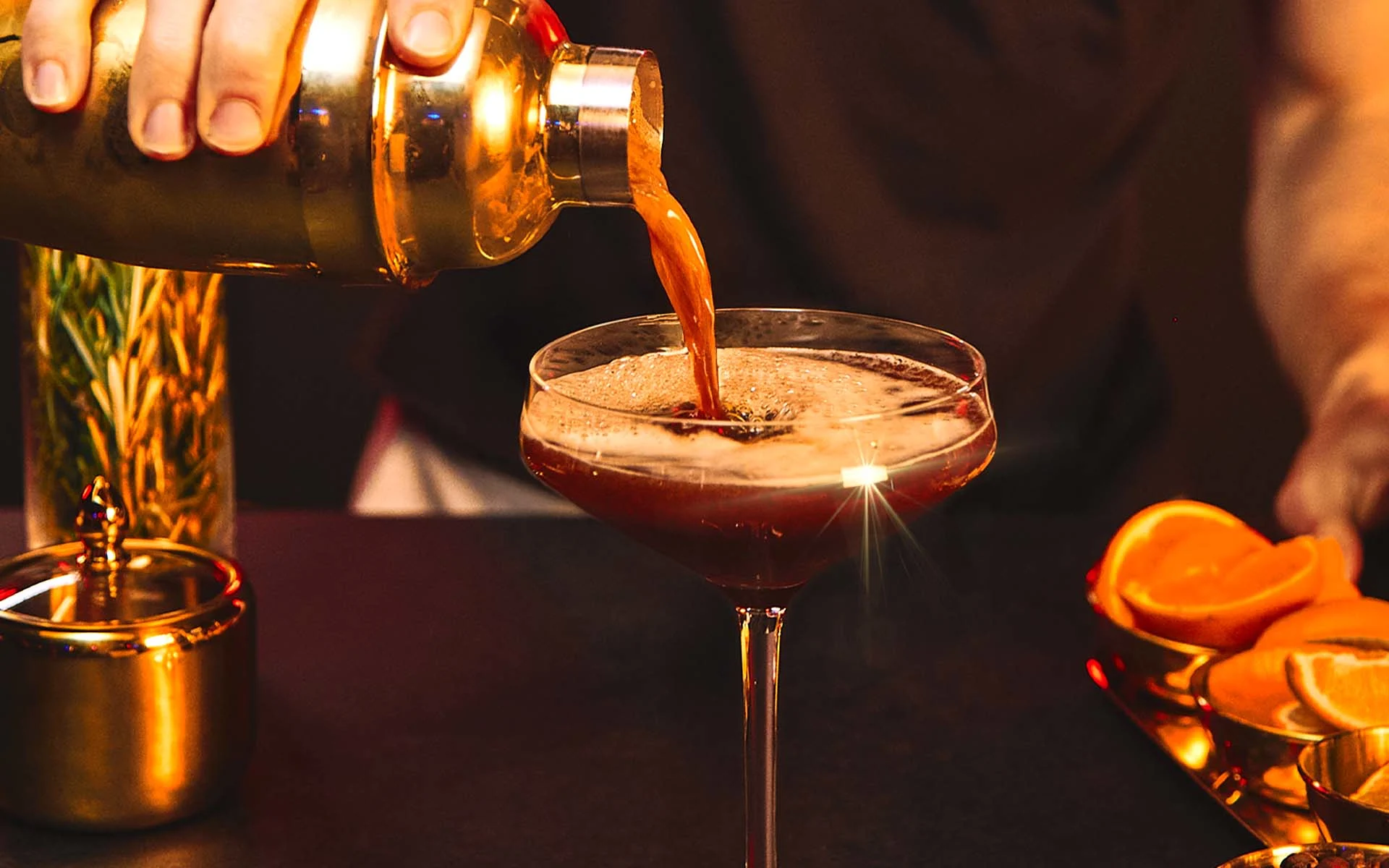 Drink being poured from a cocktail shaker into a glass with oranges on a side platter