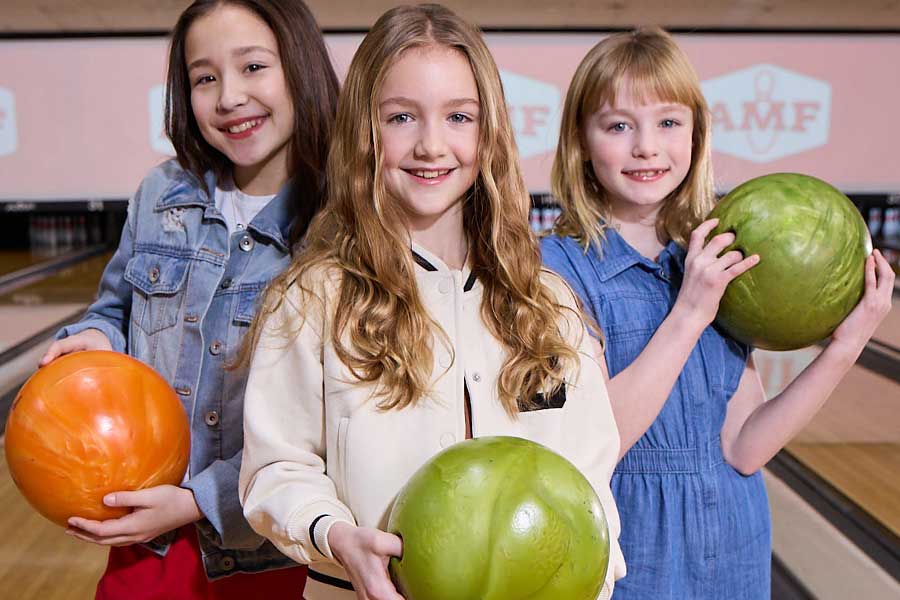 Three girls smiling and holding bowling balls while standing at the start of a bowling lane.