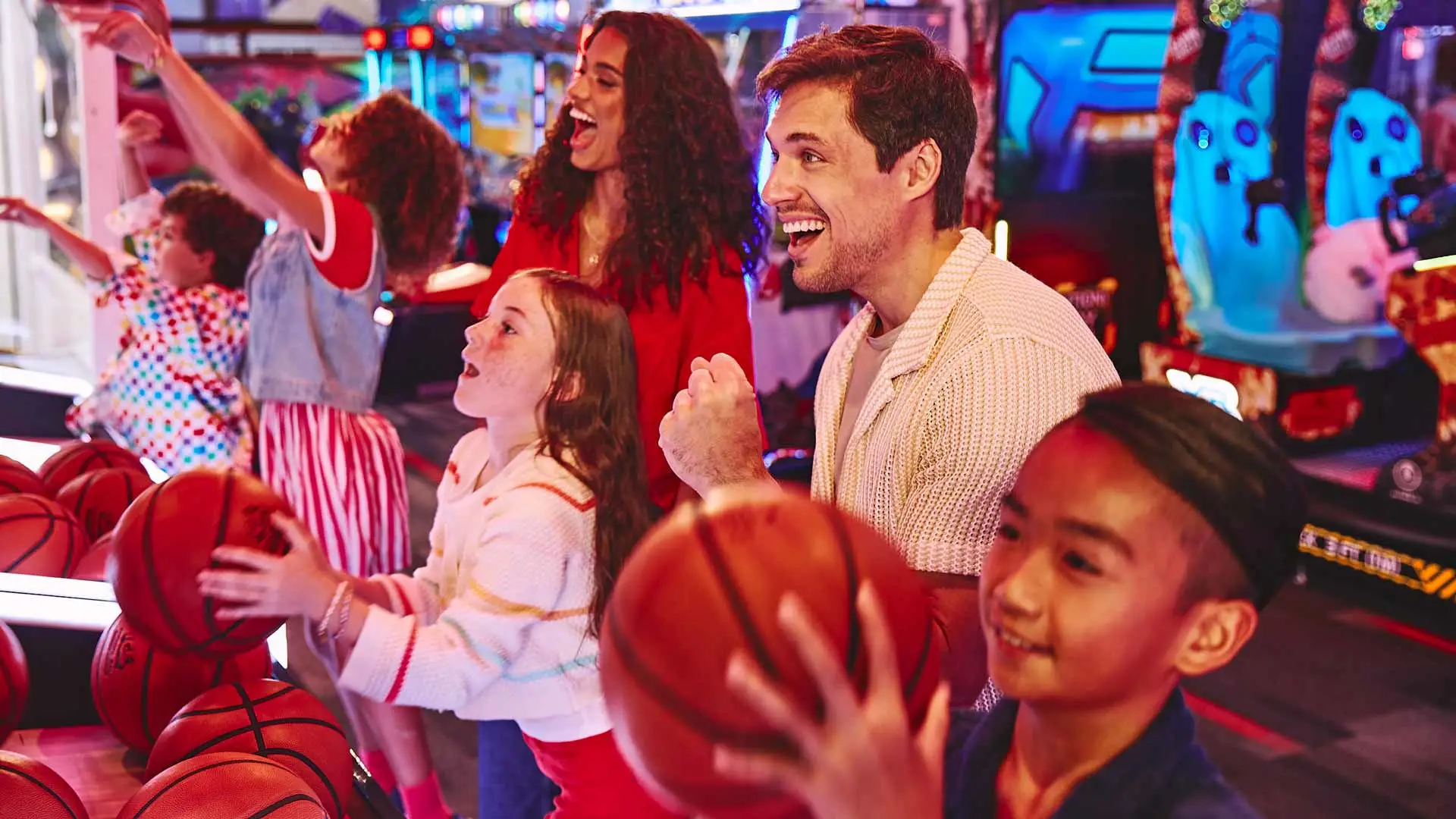 Family enjoying arcade games together, with kids holding basketballs and playing side-by-side while adults cheer them on in a colorful, neon-lit entertainment center.