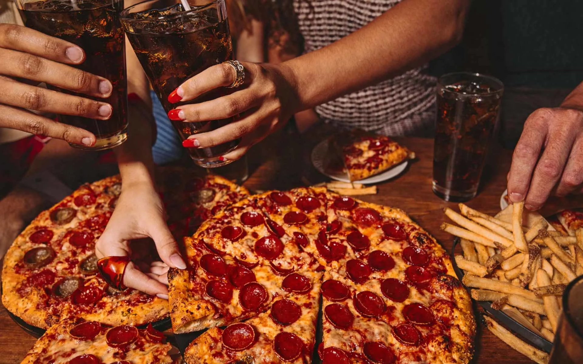 Hands toasting with glasses of cola over a table filled with pepperoni pizza and fries, capturing a lively, shared dining moment in a warm, social setting.