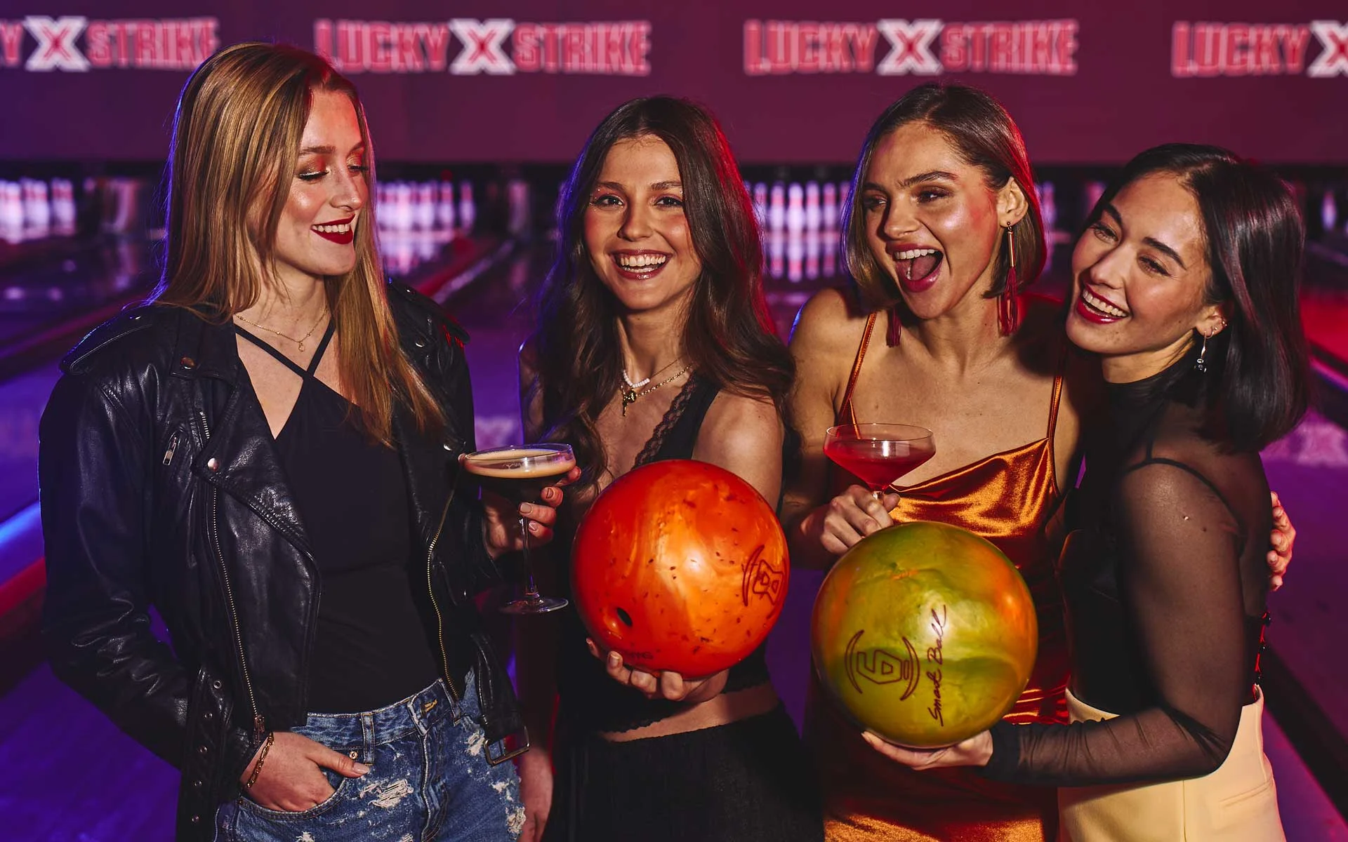 Four women enjoying a night out at a modern bowling alley, smiling and holding cocktails and bowling balls under neon lighting with lanes glowing in the background.