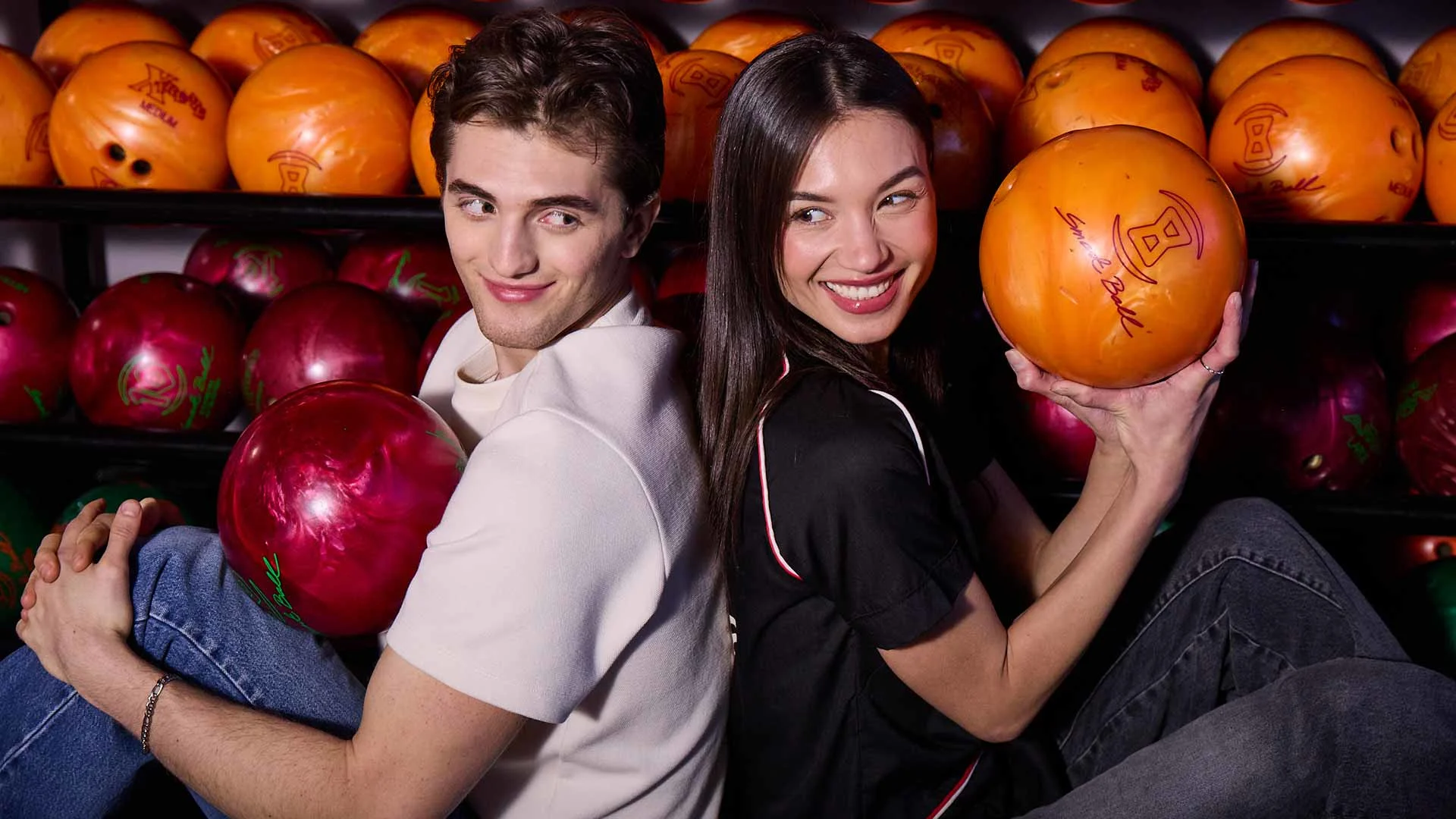 A smiling couple sitting back-to-back at a bowling alley, each holding a bowling ball in front of a rack of colorful bowling balls.