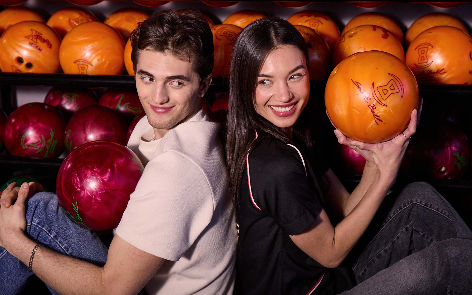A smiling couple sitting back-to-back at a bowling alley, each holding a bowling ball in front of a rack of colorful bowling balls.