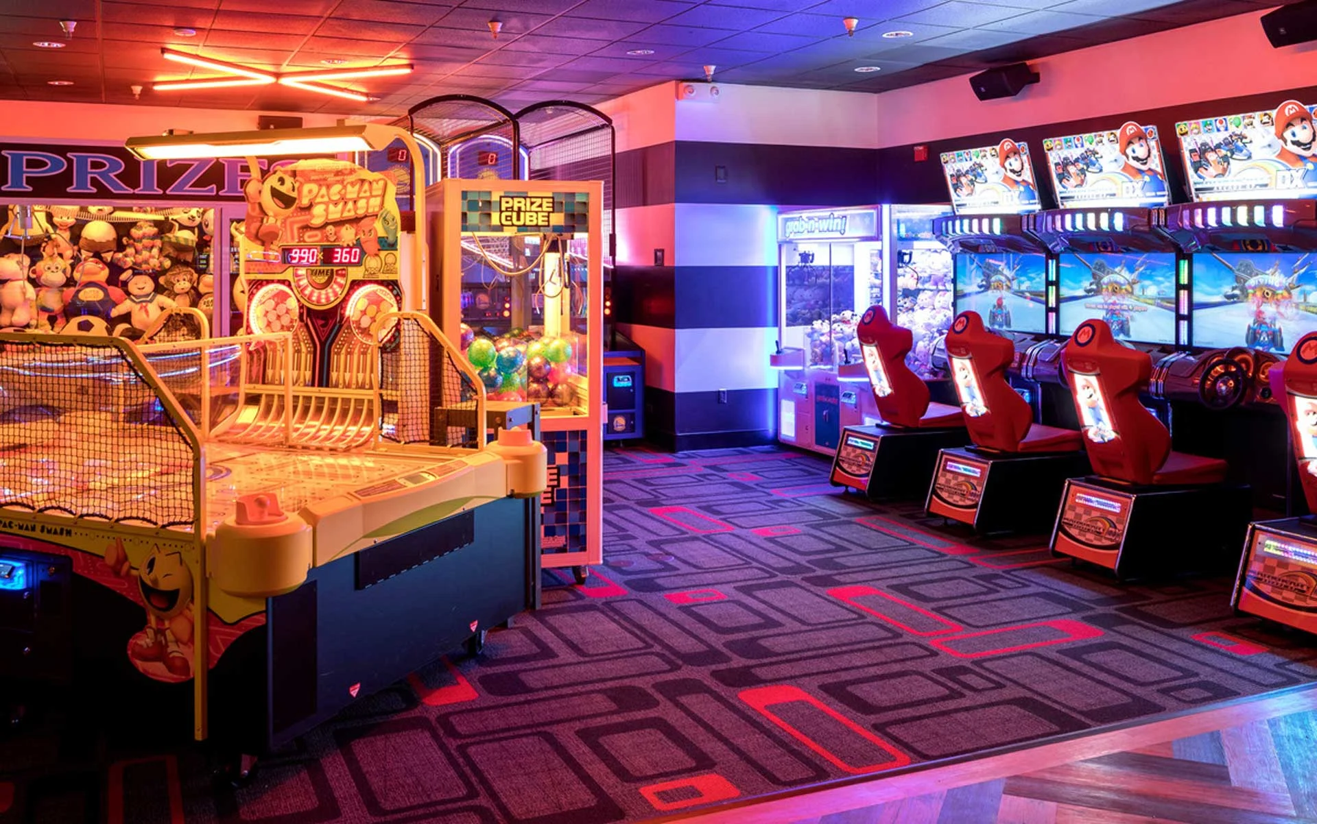 Colorful arcade area with neon lighting, featuring prize machines, skee-ball, and Mario Kart racing games, set against bold patterned carpet and a lively, family-friendly atmosphere.