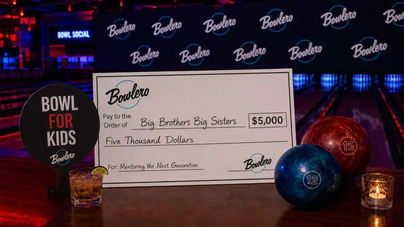 Diverse group of adults laughing at a Bowlero charity bowling event, holding bowling balls, drinks, a donation check, and a “Bowl for Kids” paddle.