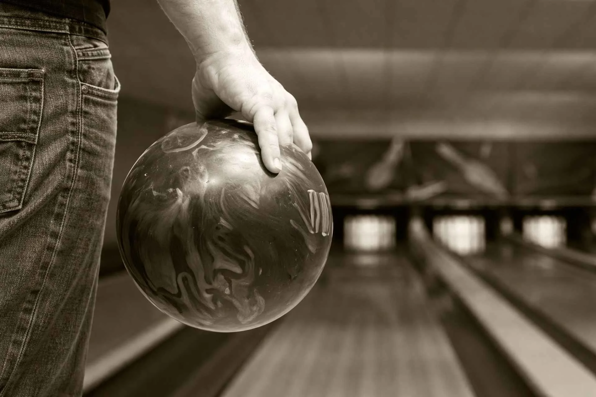 Close-up of a bowler holding a marbled bowling ball beside a lane, with pins blurred in the background.