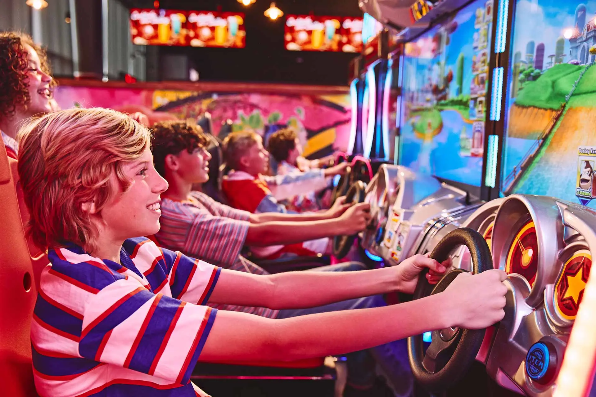 Kids playing racing arcade games side by side, smiling and gripping steering wheels in a brightly lit arcade.