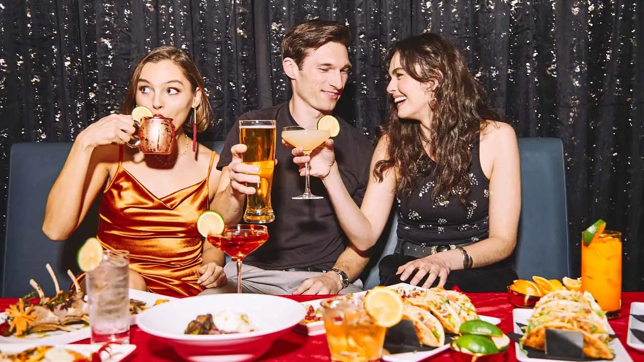 A group of two women and one man sitting in a booth lounge with a table spread of food cheersing drinks at a Lucky Strike center.