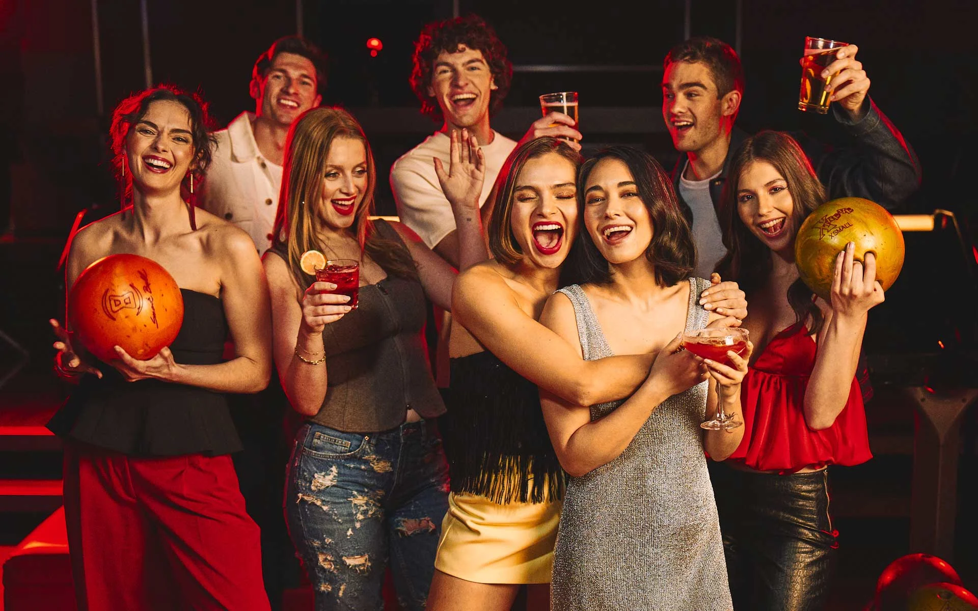 Group of young adults celebrating with drinks and bowling balls in a stylish, nightlife-inspired bowling venue with warm red lighting.