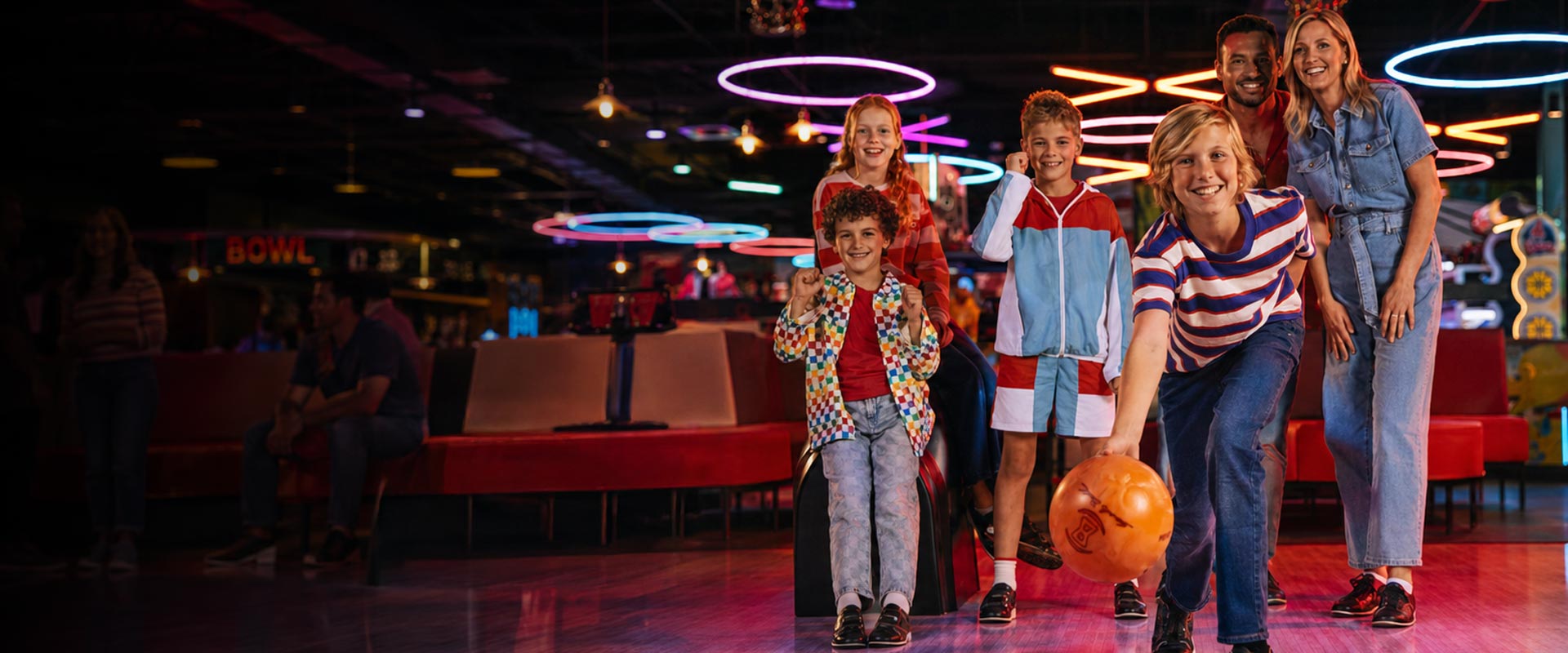 Family bowling together at a lively entertainment venue with neon lighting, arcade games, and guests enjoying food and drinks.