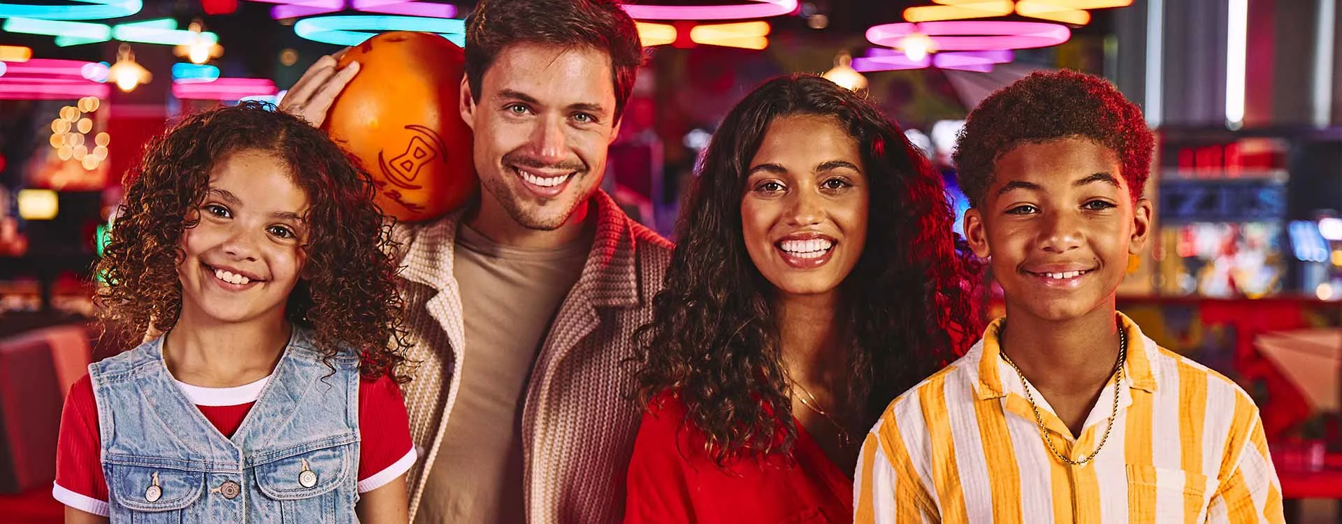 Smiling family posing together at a bowling alley with colorful neon lights and arcade games in the background.