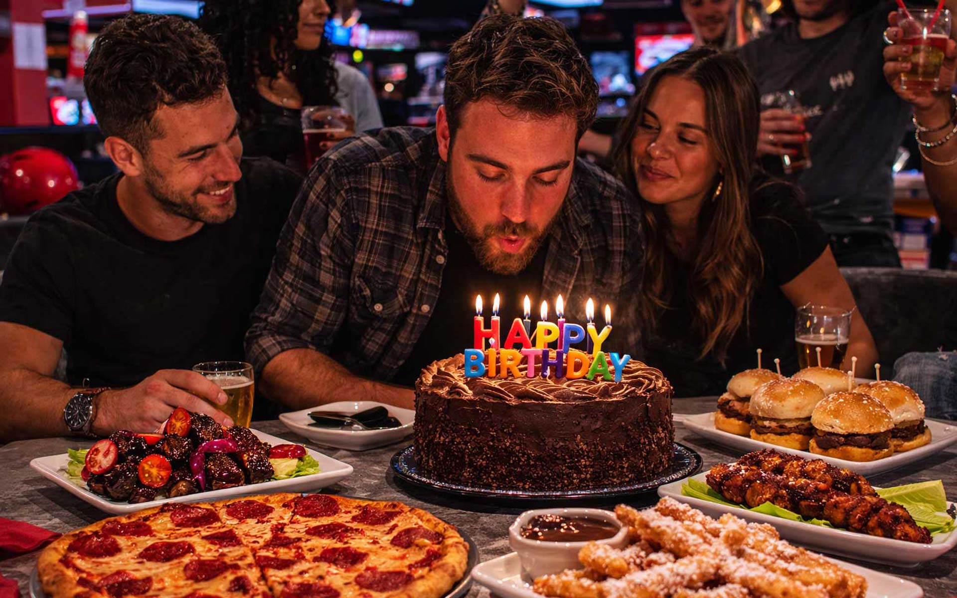 Friends celebrating a birthday at an AMF bowling center as a man blows out candles on a sprinkle-covered cake while pizza, sliders, wings, and funnel cake fries sit on the table in front of them.