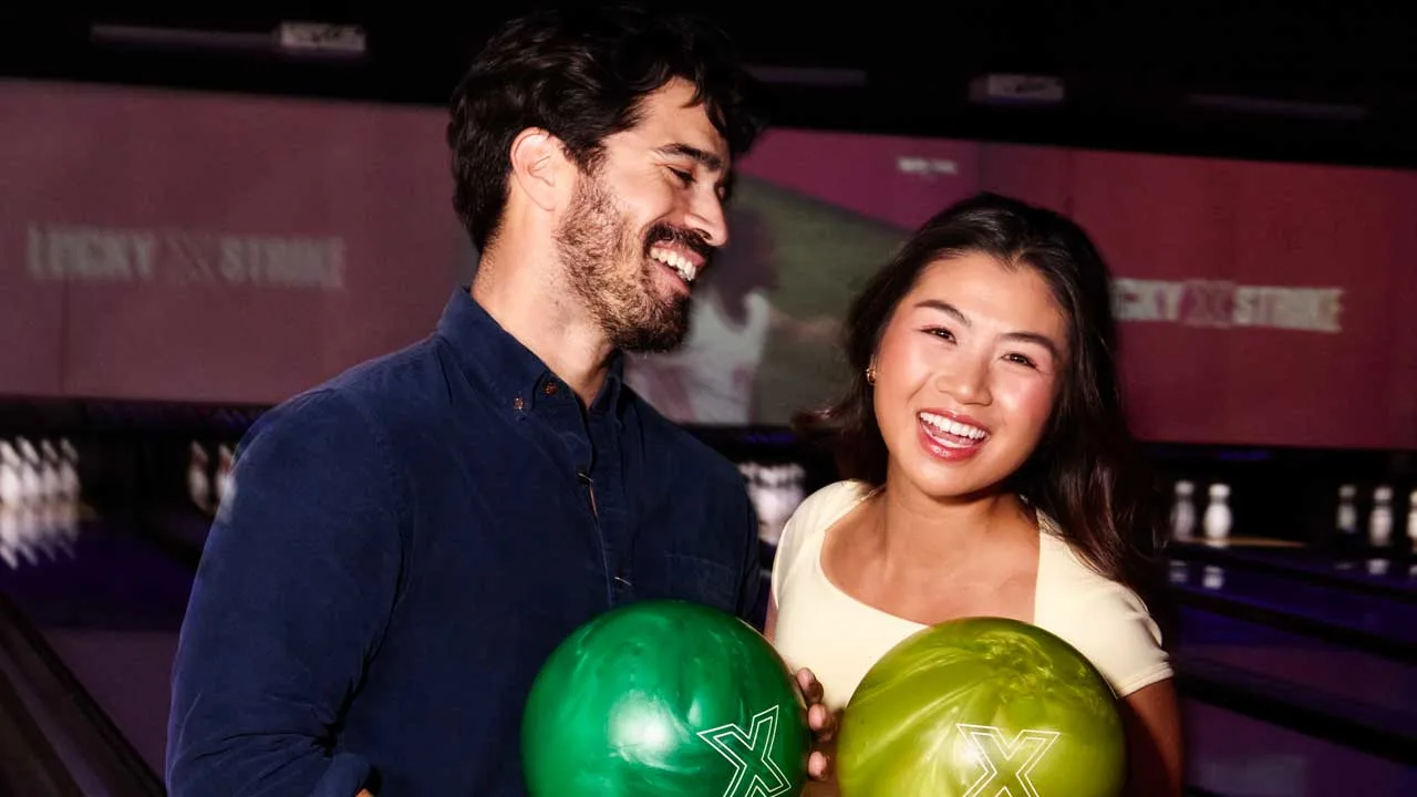 A couple standing on the approach of a bowling lane holding bowling balls smiling.